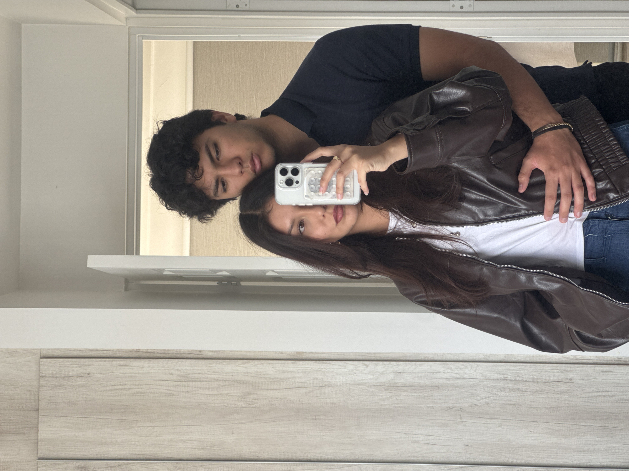 A woman is taking a selfie with a man, possibly in a residential setting. The woman is holding a phone with a patterned case, wearing a brown leather jacket over a white t-shirt and jeans. The man is behind her, wearing a black shirt. They are positioned in front of a mirror with a light-colored wall and a wooden surface below, likely a cabinet or panel. The setting appears to be indoors. There is no information to confirm they are in San Francisco Ocotlán, Mexico.