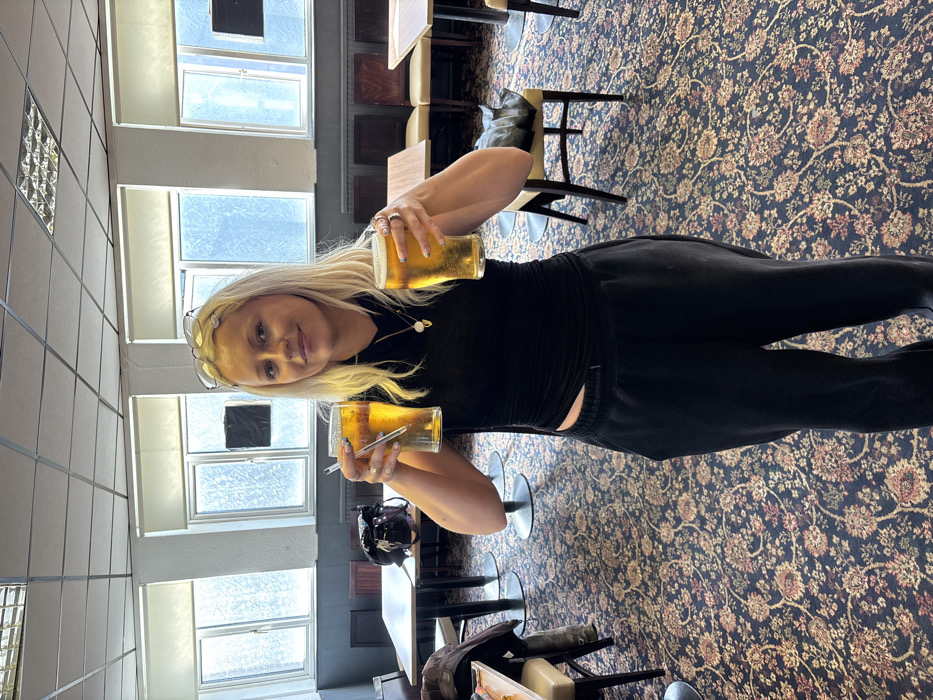 A female subject stands centrally within a public house or bar interior in London, United Kingdom. She is an adult with blonde hair, wearing glasses on her head, a black long-sleeved top with a waist cut-out, black sweatpants, and two necklaces. She holds a glass of amber liquid, consistent with lager or cider, in each hand, extended towards the viewer; a straw is present in the glass in her left hand (viewer's right). A septum piercing is visible. The floor is covered by a dark, multi-colored floral patterned carpet. The background features multiple tables with light-colored tops and dark bases, and various chairs, some upholstered in beige. A black bag is on a chair to the left, and a dark jacket drapes over a chair to the right. Three large windows illuminate the scene, with radiators below them, and the view outside is indistinct. The walls beneath the windows are dark, and the upper walls are white. The ceiling is a grid-style suspended ceiling with recessed lighting.