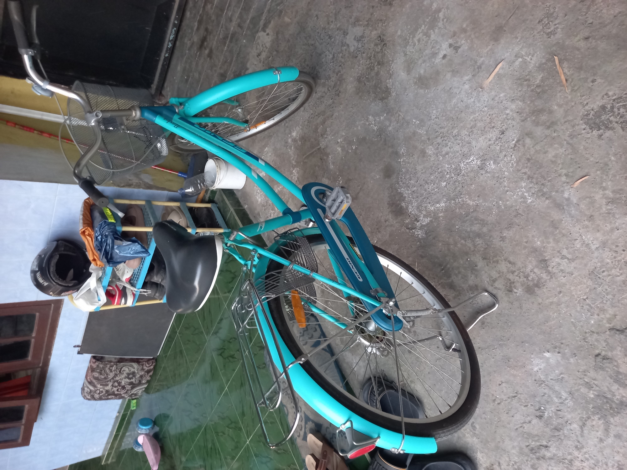A teal bicycle is parked on a concrete floor in front of a blue-walled building in Kebon Agung, Indonesia. The bicycle has a front basket, a rear rack, and a kickstand. There are various objects scattered around the scene, including a black helmet, shoes, a plastic toy, and a white bucket with a bottle of water. The floor is tiled in a green diamond pattern near the building and transitions to rough concrete. The lighting suggests it is daytime, with shadows indicating the sun is at an angle.
