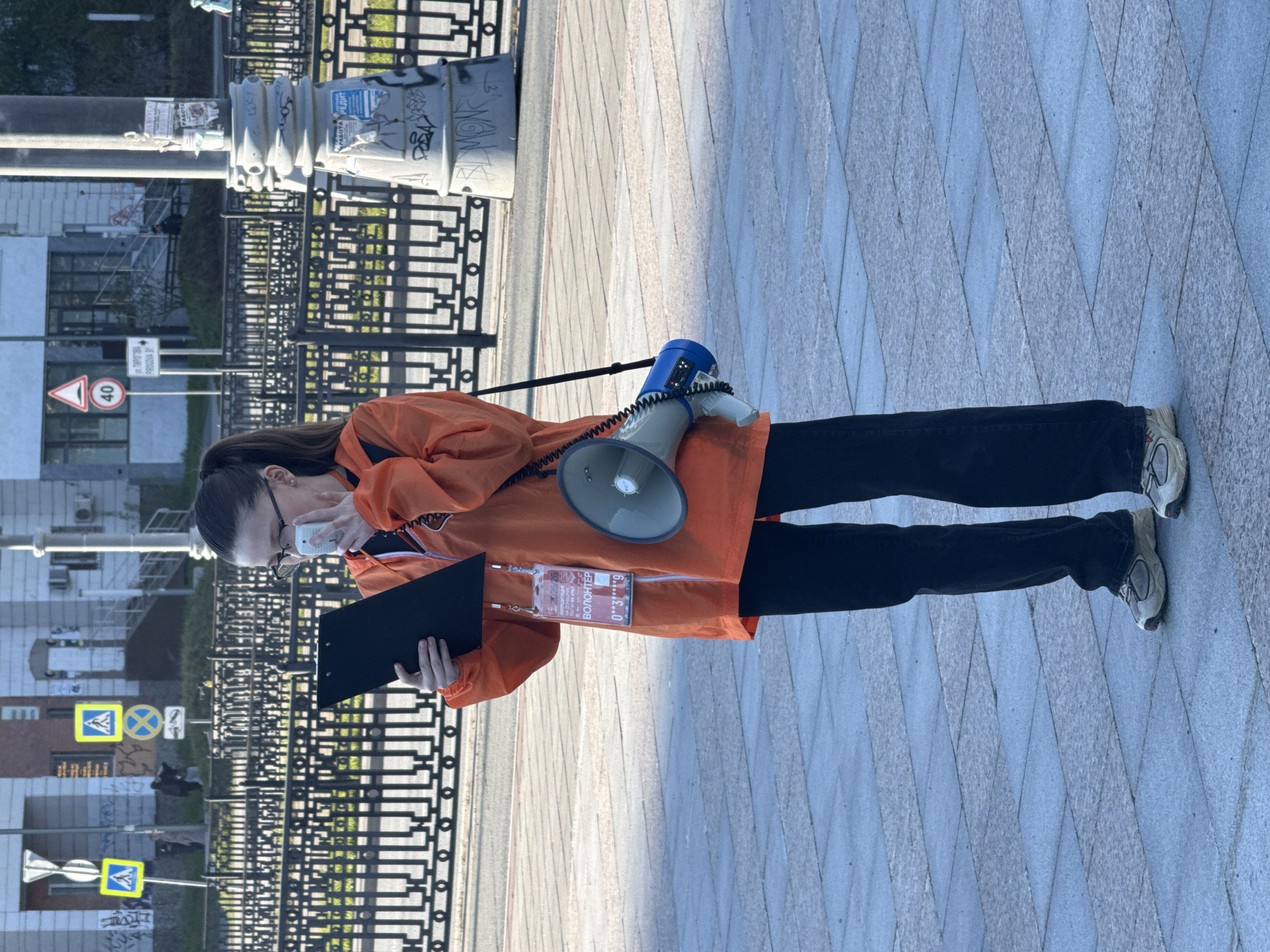 A person in an orange jacket and black pants stands on a sidewalk of rhomboid tiles in Yekaterinburg, Russia. The person is holding a black clipboard in their left hand and talking on a white cell phone held to their ear with their right hand. A blue and gray megaphone hangs from their shoulder. A lanyard with a badge that reads "ВОЛОНТЕР" (Volunteer) hangs around their neck. A black metal fence is visible to the left, separating the sidewalk from a street with pedestrian crossing signs. Behind the fence are buildings and a lamp post covered in graffiti. The weather is sunny, creating shadows on the sidewalk.