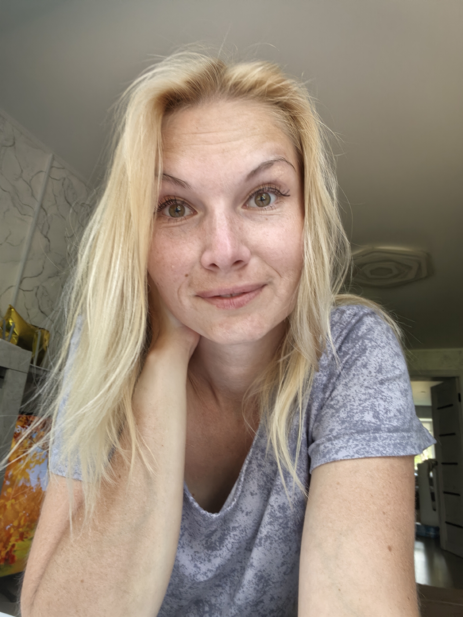 A woman with blonde hair and light skin is looking at the camera. She is wearing a grey t-shirt. Her left arm is bent and her hand is resting on her neck. She has freckles on her face and arm. The background is blurred, but it appears to be an indoor setting. The image was taken in Sterlitamak, Russia.