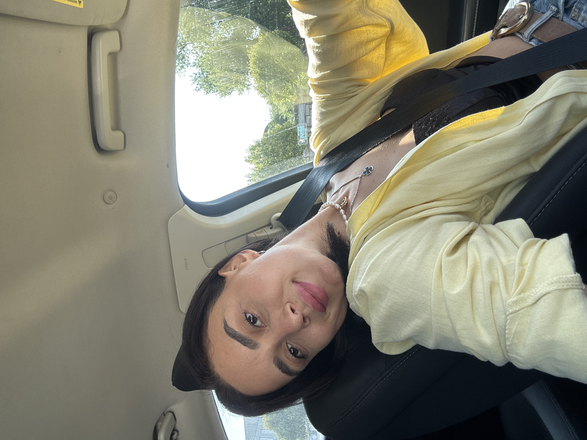 A young woman with dark hair, wearing a yellow cardigan over a black top and a pearl necklace with a cross pendant, is seated in the passenger seat of a car. She is leaning her head to the side and smiling at the camera. The interior of the car is visible, including a black seat, a seatbelt, and the car's ceiling with an airbag label. Outside the car window, trees and a building are visible, suggesting an urban environment. A sign with text reflected in the window indicates the location is Tbilisi, Georgia.