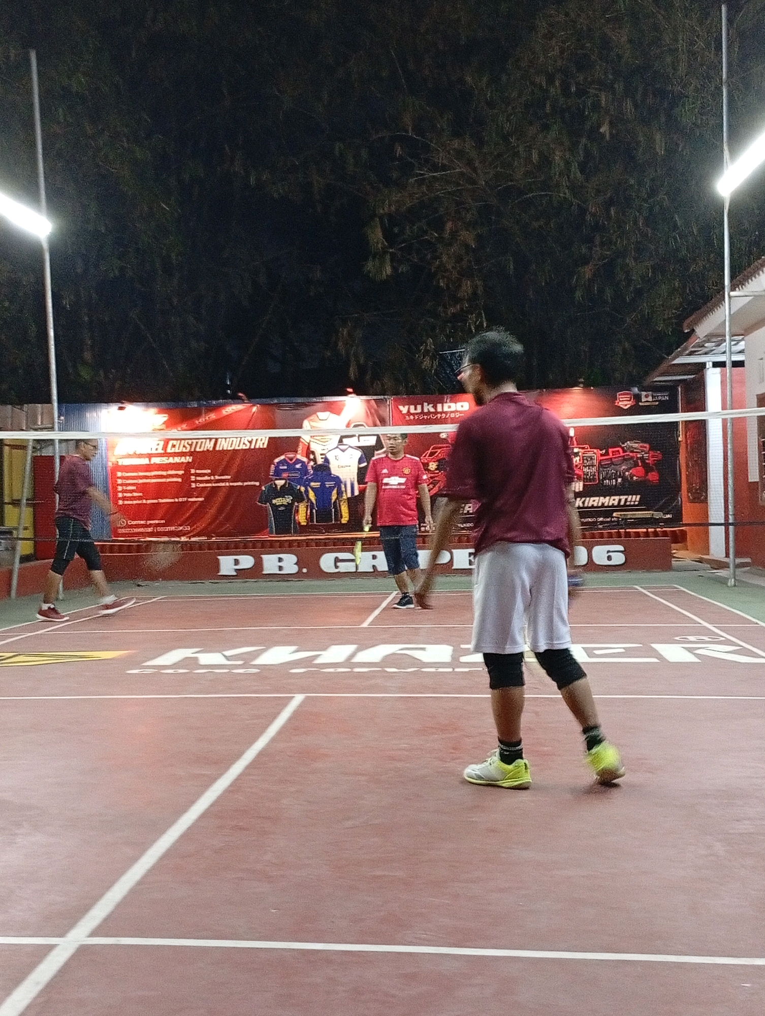 The image captures a nighttime outdoor badminton game in progress on a red court marked with white lines. Two men are visible on the court, engaged in play. One man, wearing a maroon t-shirt and black shorts with knee pads, is in the foreground, facing away from the camera, with a badminton racket in his right hand and a yellow shuttlecock near his left foot, indicating he is about to strike it or has just done so. Another man, wearing a red t-shirt and denim shorts, is on the other side of the net, also with a racket. In the background, behind the net, a large red banner with white lettering advertises "PB. GARUDA 06" and other text, likely indicating the name and possibly the location of the badminton club. The court is illuminated by bright overhead lights, casting shadows on the court. Trees and a building are visible in the dark background. The scene is likely located at a badminton court in Caringin, Indonesia, suggested by the presence of "PB. GARUDA 06" which likely stands for Persatuan Badminton Garuda 06, a common naming convention for sports clubs in Indonesia.