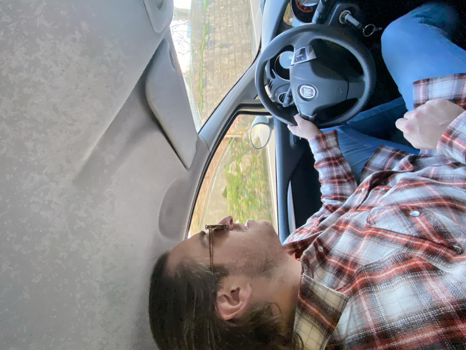 Inside a car, a man in a plaid shirt holds the steering wheel, his head tilted back against the ceiling. He wears glasses and has a beard. Outside the window, trees and buildings are visible. The interior of the car features a gray ceiling and a side mirror. The steering wheel has the Fiat logo. He is wearing blue jeans.