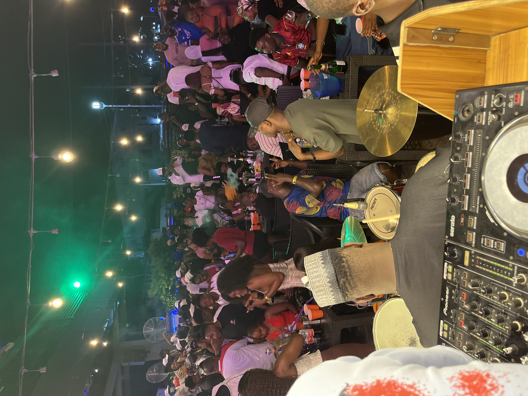 An outdoor evening gathering is underway at an unknown location. A crowd of people are socializing and enjoying themselves. In the foreground, a DJ is visible behind a Pioneer Serato DJ mixer, with drum cymbals and parts of a drum kit to his left and right. The DJ is wearing a black shirt and a patterned cap. In the middle ground, a live musical performance is taking place, with a drummer playing a Premier drum and a male performer singing into a microphone. Various people are seated at tables, with drinks and hookahs present. The background shows more people dancing and mingling, with building structures and streetlights visible in the distance. The scene is illuminated by string lights and green spotlights.