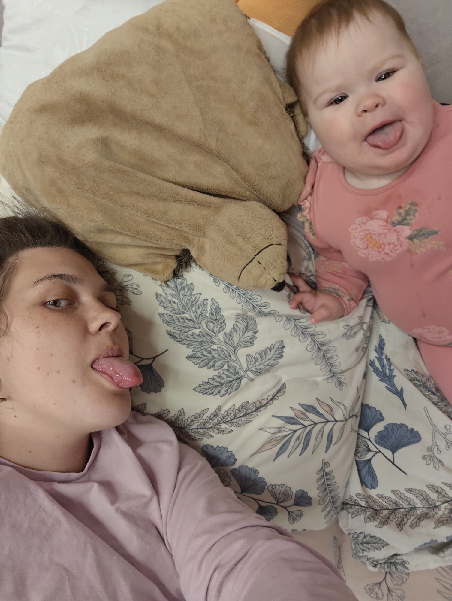This intimate indoor photograph captures a heartwarming and playful moment between a woman and a baby, taken in Chaikovsky, Russia. Both individuals are lying down, appearing to be on a bed, and are playfully sticking their tongues out towards the camera, suggesting a shared, lighthearted interaction.
The woman, positioned on the left side of the frame, has light brown hair, visible freckles on her face, and is wearing a light purple long-sleeved shirt. Her left eye is visible, looking towards the right, matching the baby's playful expression.
The baby, located on the right, has light brown hair and rosy cheeks. The baby wears a pink long-sleeved outfit adorned with a floral pattern featuring pink, white, and green blossoms. A large, light brown plush teddy bear or soft toy is visible between the two, partially behind the baby's head and neck.
The bedding beneath them is a light-colored sheet with a distinctive pattern of blue and grey leaves, including ginkgo-like shapes. A plain white pillow is partially visible behind the woman's head. The overall mood of the image is joyful and affectionate, capturing a candid and fun interaction. There is no visible text in the image.