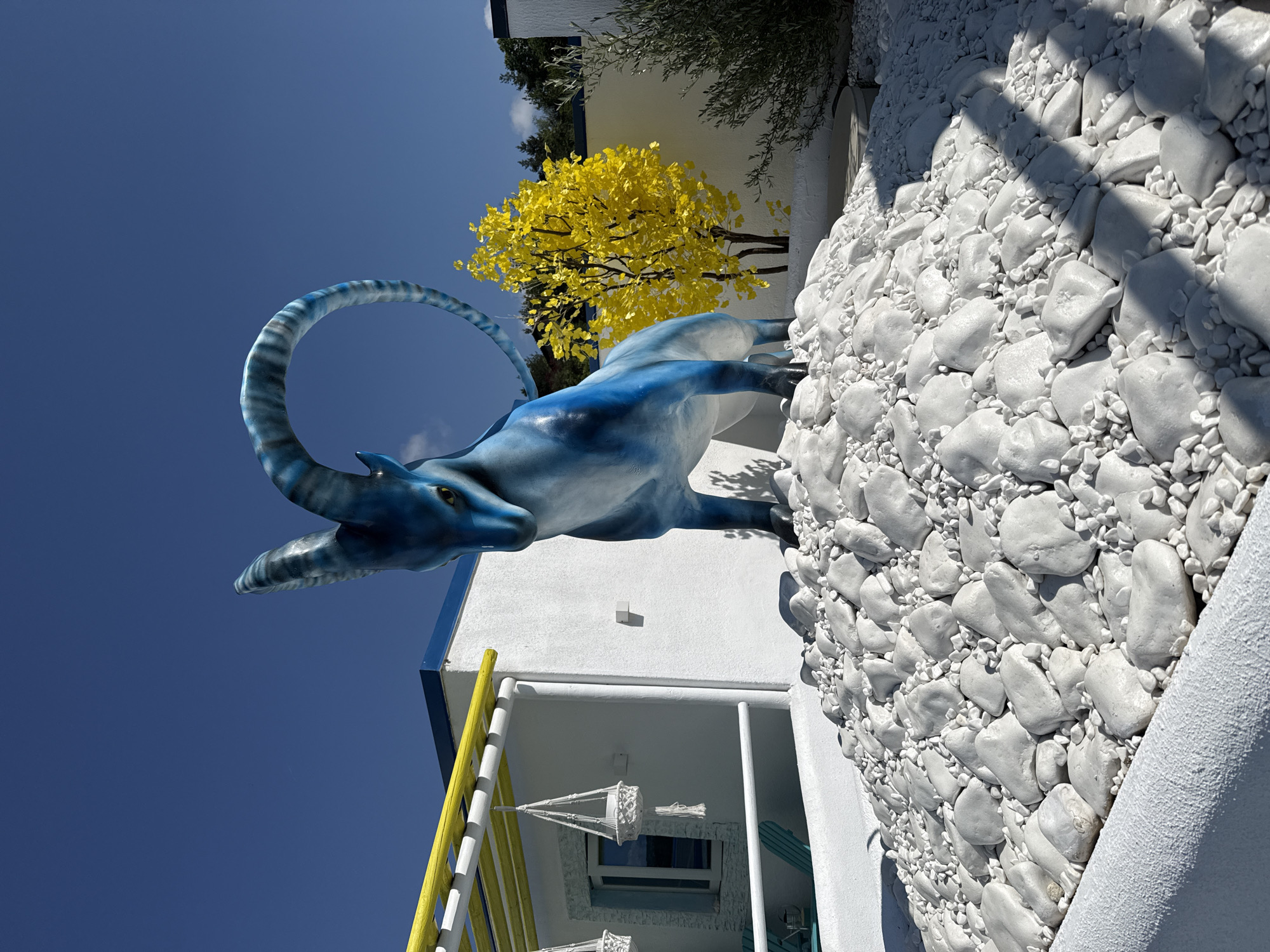 In Baliovtsi, Bulgaria, a striking blue statue of an ibex with spiraling horns is positioned prominently in an outdoor setting. The statue stands on a mound of white rocks and pebbles, creating a stark contrast against the vibrant blue sky. To the left of the statue, a white building with a yellow-trimmed pergola is visible. Hanging from the pergola are two white macrame plant hangers. In the background, a small tree with bright yellow foliage stands out, and to its right, a lush green shrub or small tree is present. The area is illuminated by bright sunlight, casting shadows on the white rocks. The overall scene depicts an artistic and somewhat whimsical outdoor display.