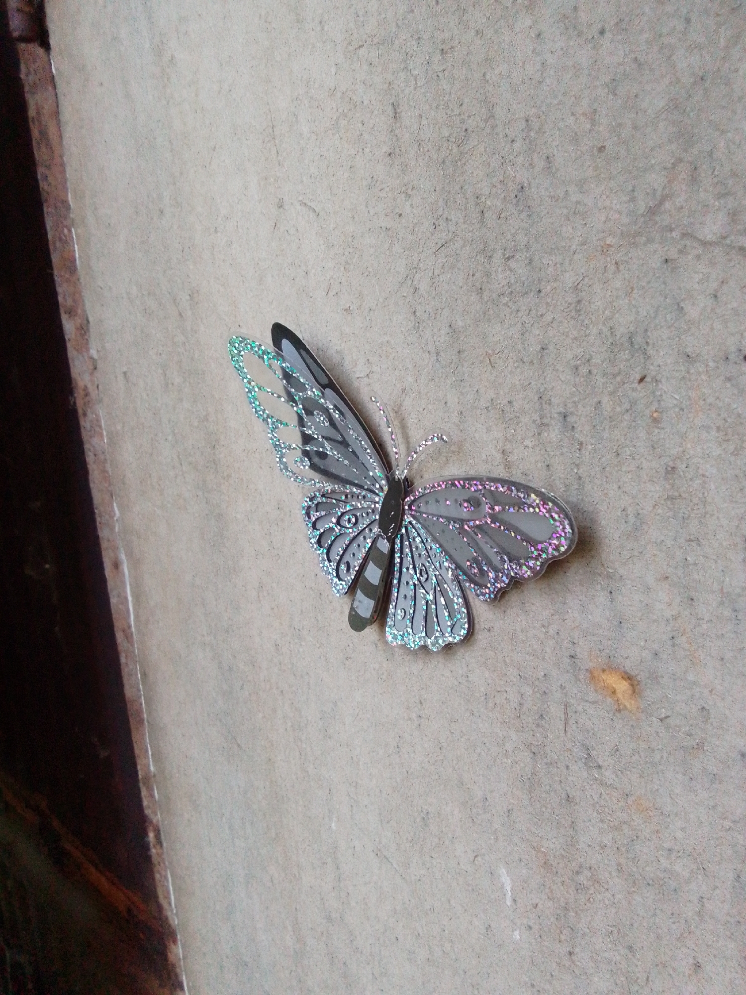 A decorative butterfly pin with gray and iridescent wings is affixed to a textured, light-colored concrete wall. The butterfly has intricate patterns on its wings, highlighted by sparkling accents. To the left of the wall, a dark, reddish-brown metallic surface is visible, possibly a metal beam or frame. The environment appears to be outdoors, with natural light illuminating the scene. No people are present, and no specific location within Pavlodar, Kazakhstan, can be determined from the image.