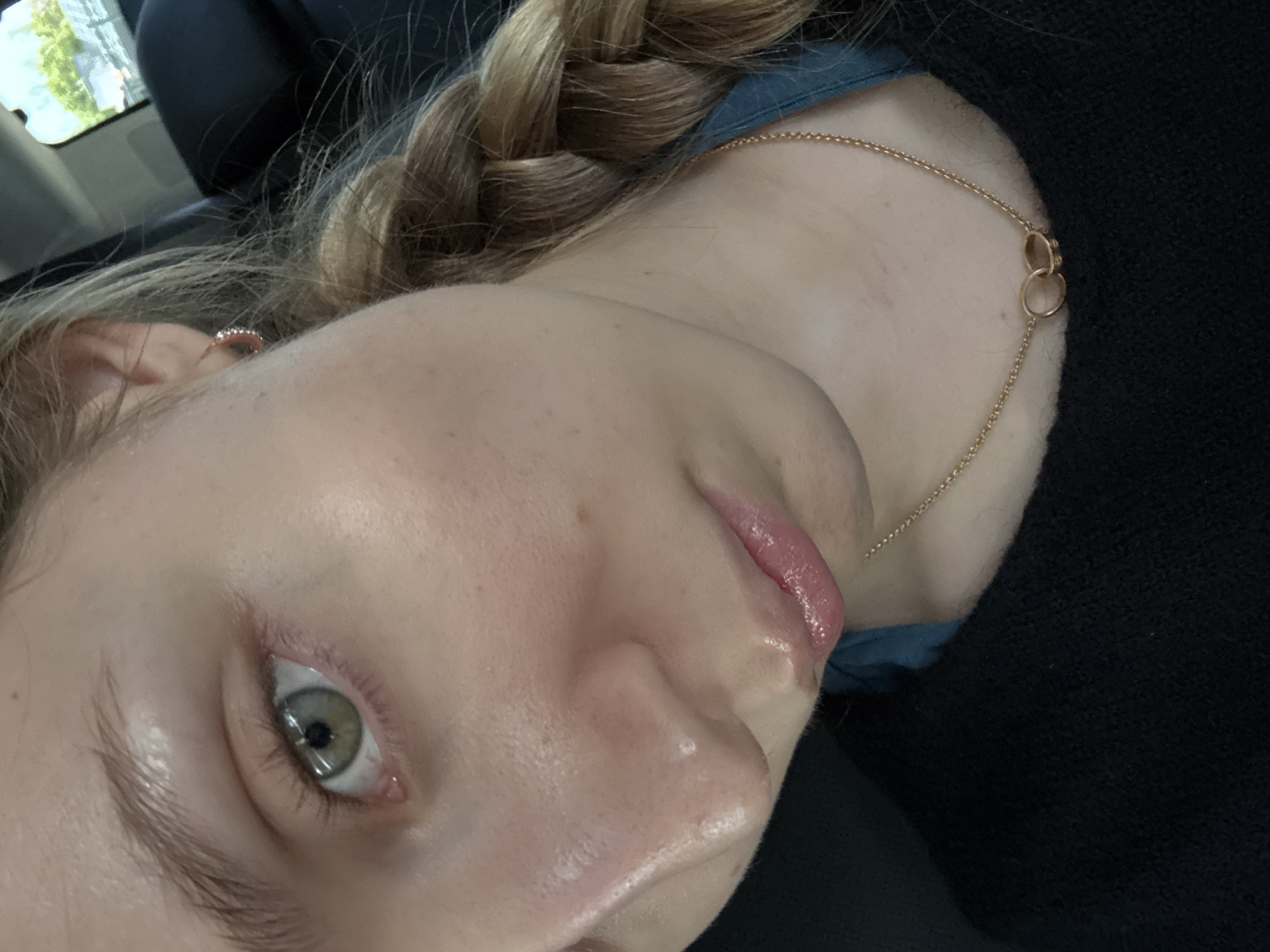 A close-up, tilted portrait captures the upper body of a fair-skinned person, likely a young female, situated inside a vehicle in Mexico City, Mexico. The person's head is angled, with their left eye looking directly towards the camera, revealing a green or hazel iris with a dilated pupil. Their lips are glossy and pink.
The subject's light brown or blonde hair is styled in a thick braid, visible on the left side of the frame. A small, delicate earring with what appears to be tiny stones is worn in the left earlobe. Around their neck, a thin gold chain necklace with two interlocking gold rings is visible, resting against the skin just above a blue garment and a dark, possibly black, textured sweater or fabric covering the shoulder. The person's skin shows a few small freckles or moles.
In the blurred background, through what appears to be a car window, faint hints of green foliage and distant buildings suggest an outdoor, urban environment during daylight hours. No discernible text is visible in the image.
