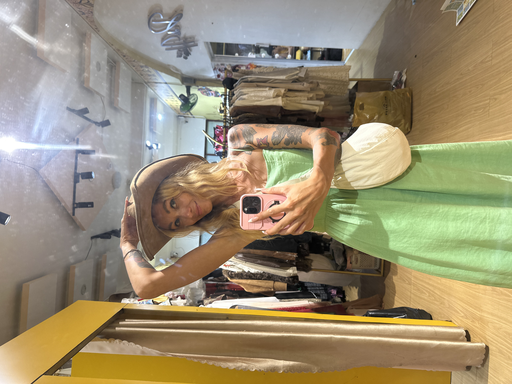 In Phu Thuy Ward, Vietnam, a young Caucasian woman with blonde hair, wearing a light green sundress and a brown cowboy hat, is taking a mirror selfie in a boutique. She holds a pink phone in her right hand. She has tattoos on her left arm and chest. A light-colored purse is slung over her shoulder. The background shows racks of clothing, a yellow changing booth, and a reflective ceiling. The floor is wooden.