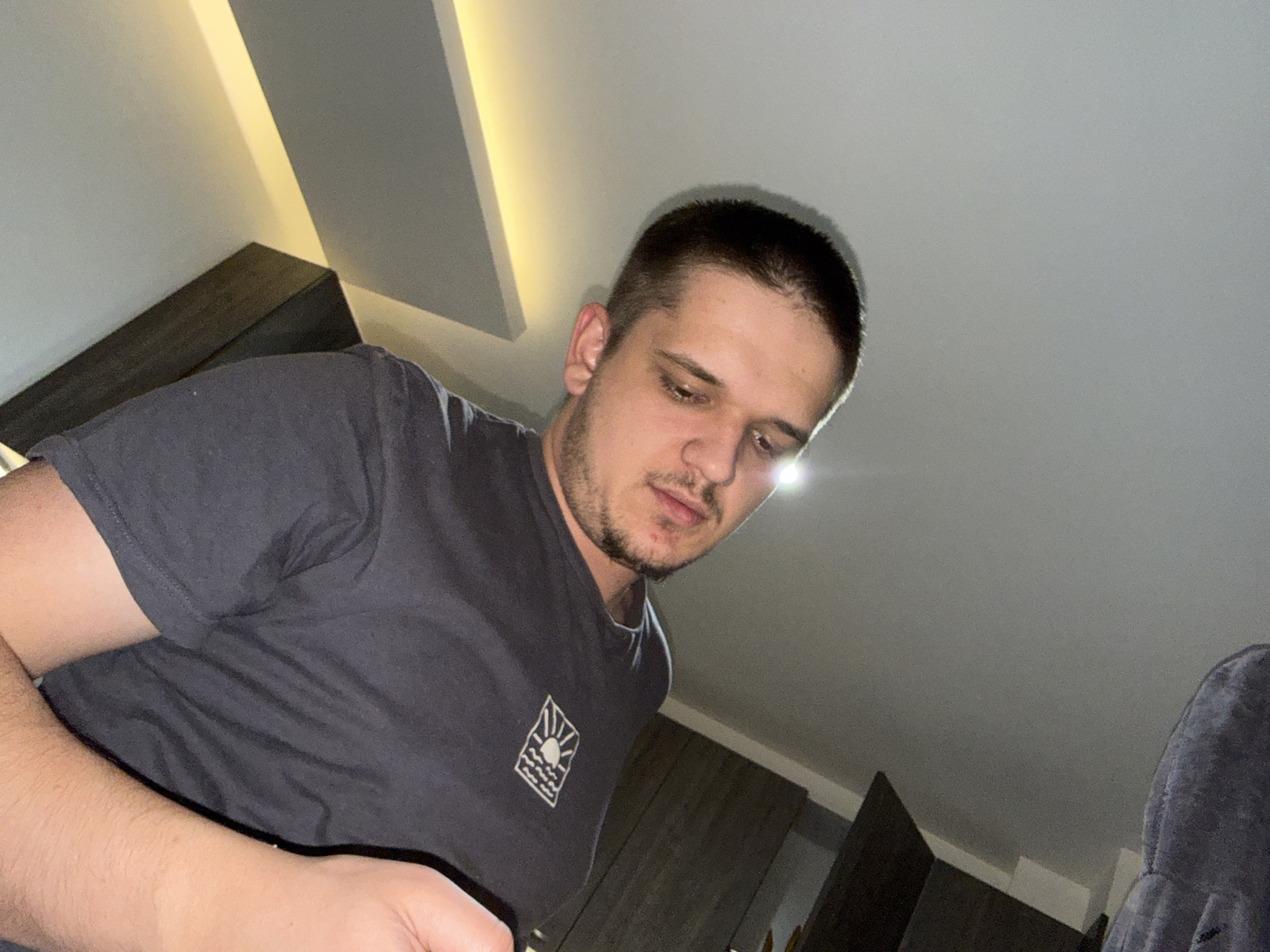 A man is captured in a low-angle, close-up shot indoors. He has short dark hair, stubble on his face, and is wearing a grey t-shirt with a small logo on the chest. The environment includes a ceiling with what appears to be recessed lighting. Behind the man, there are dark-colored cabinets or shelves. The location is unknown.