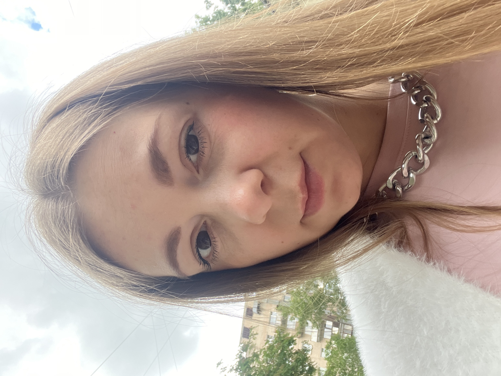 A close-up, angled selfie captures a young woman with long, straight blonde to light brown hair framing her face. She has light-toned skin, natural eyebrows, and dark eyelashes, and looks directly at the camera with a neutral expression and slightly parted lips. She is wearing a light pink top or dress, accessorized with a prominent, chunky silver chain-link necklace. A fluffy white garment, possibly a sweater or jacket, is also visible over her shoulder or in the foreground.
The background shows an outdoor setting with green trees and a light-colored building, likely an apartment block or similar urban structure, featuring multiple windows. The sky overhead is overcast, indicating daytime. The image is composed as a selfie, with the subject's face dominating the frame and the background slightly out of focus. There is no visible text in the image.