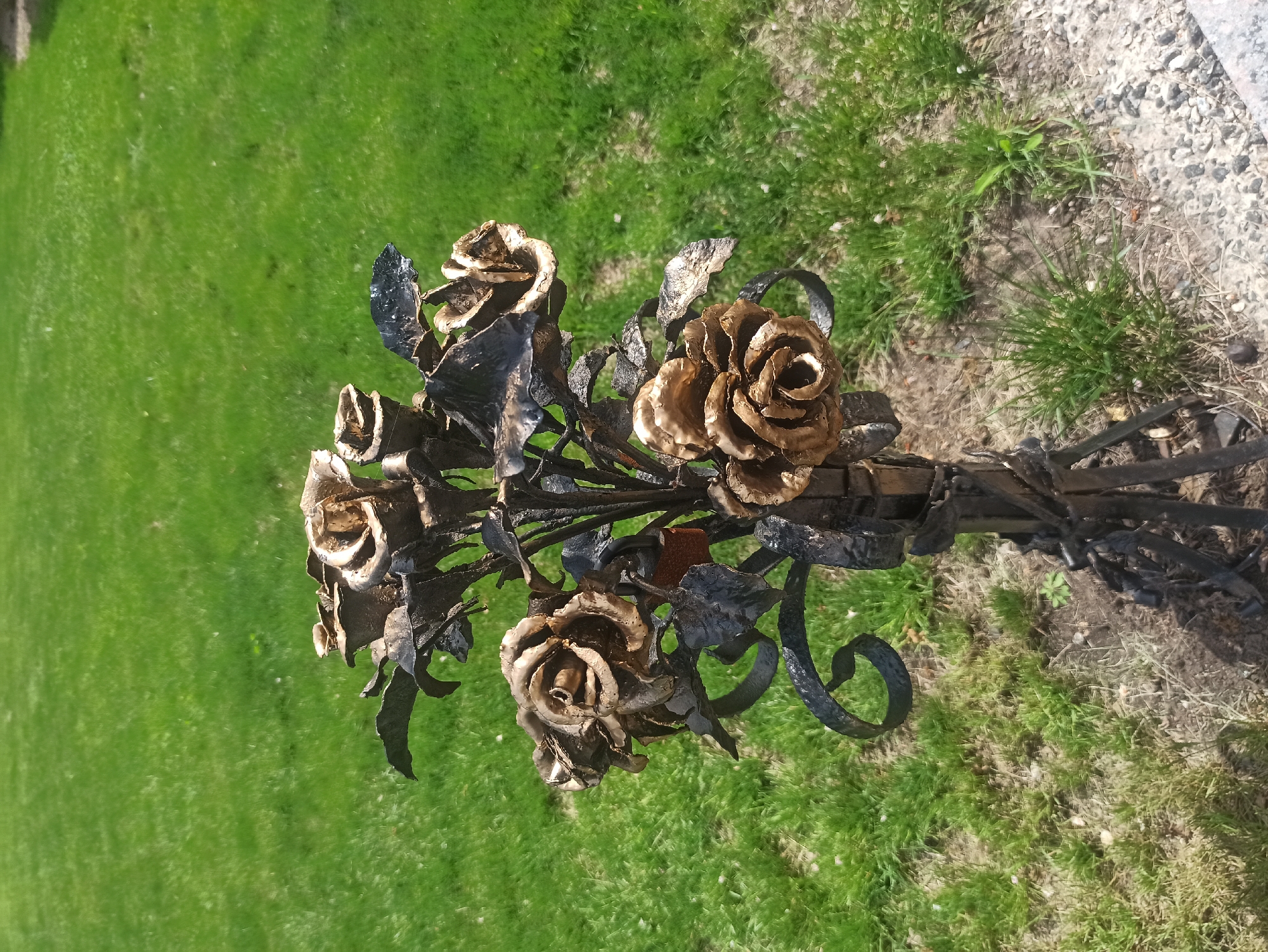 A cluster of metal roses stands on a lawn in an unidentified location. The roses are crafted from a dark metal and given a bronze-like finish on the petals. They have distinct leaves and stylized, curling stems. The scene is outdoors. The surface around the flowers is green grass. Near the base, there is gravel and some ground cover. The overall lighting suggests it is daytime. No other people or objects are visible in the image.