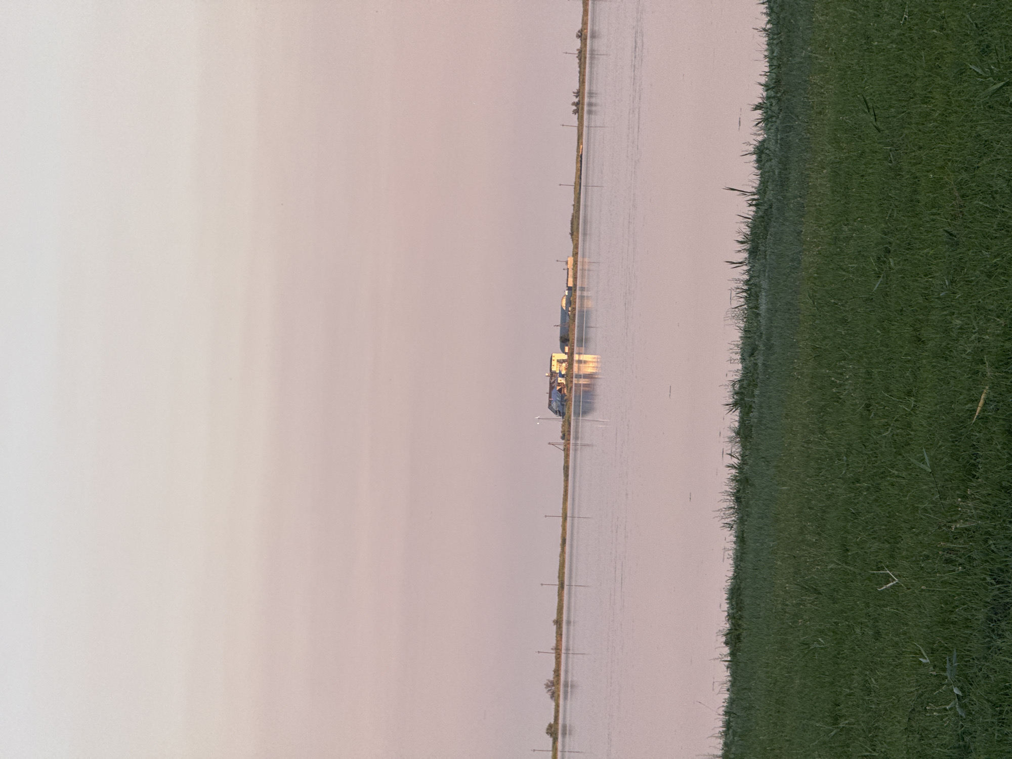 The image depicts a serene waterscape near Serhiivka, Ukraine, likely during twilight or early morning given the soft, pastel hues of the sky. In the foreground, lush green grass borders a calm body of water that reflects the gentle colors of the sky. Across the water, a strip of land is visible, sparsely dotted with vegetation and utility poles. On this land, a cluster of buildings, possibly residential or agricultural, stands out, their reflections shimmering on the water's surface. The overall impression is one of peacefulness and rural tranquility, characteristic of the landscape in and around Serhiivka.