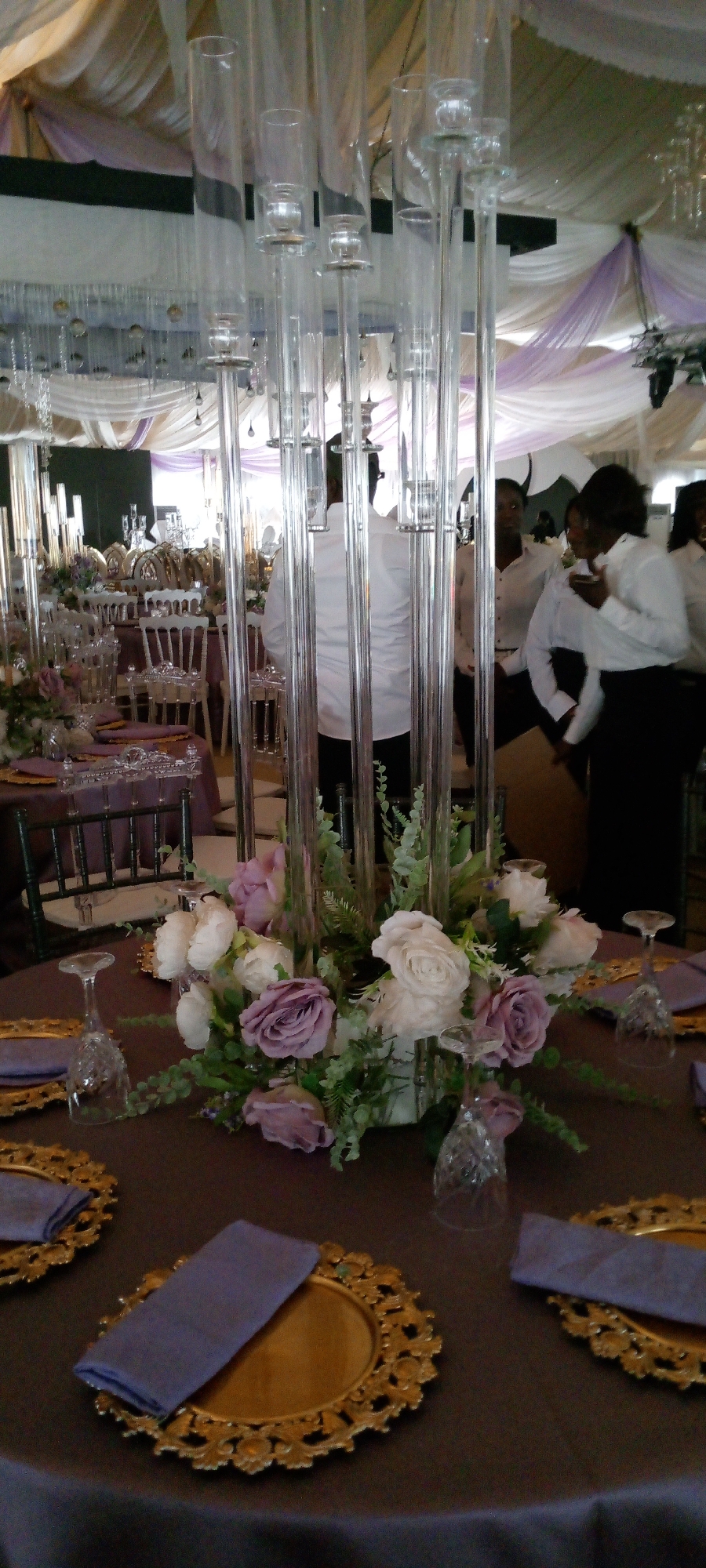 This is a medium shot, eye-level interior photograph of a formal event venue, likely a banquet hall. The foreground is dominated by a round table covered with a dark brown tablecloth. On the table, a floral arrangement featuring white and purple roses with greenery is centrally placed within a tall, multi-tube clear acrylic centerpiece. Several ornate gold charger plates with folded purple napkins are visible on the table, along with empty crystal wine glasses.
In the middle ground, more tables with similar settings, including chairs with clear acrylic accents, are arranged in rows. The room is decorated with draped fabric in shades of white and lavender, suspended from the ceiling.
In the background, several people, predominantly women wearing white shirts and dark skirts or pants, are present. Some are standing and appear to be conversing. The lighting suggests an indoor event, likely during the day or with artificial lighting. The overall impression is one of a wedding reception or a similar celebratory gathering.