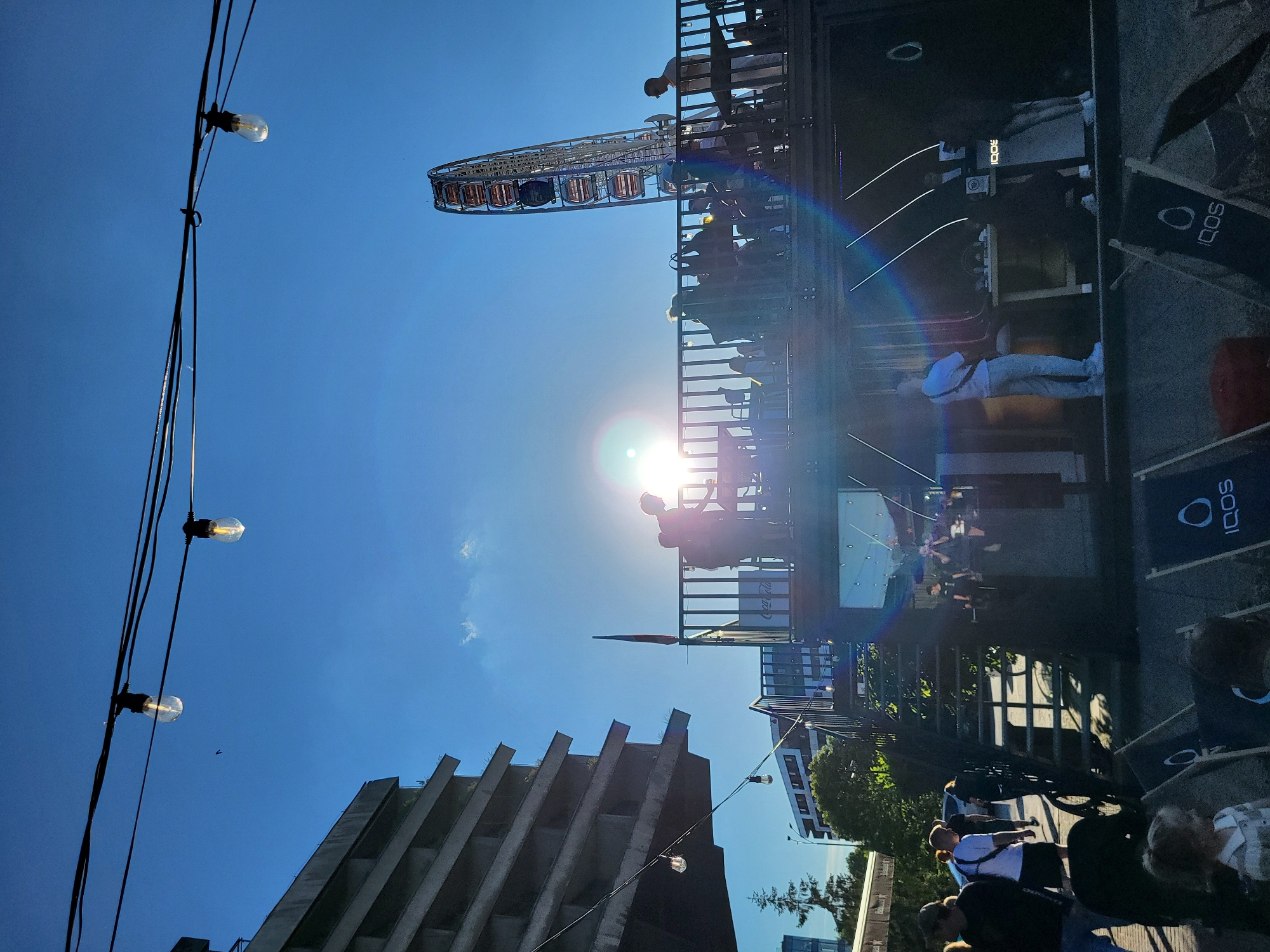 An outdoor urban scene is captured under a clear, bright blue sky with the sun positioned centrally, causing a prominent multi-colored lens flare. Overhead, two strings of decorative incandescent light bulbs are stretched across the upper frame. In the far distance on the left, a large, multi-story brutalist-style concrete building with stacked, cantilevered sections is visible.
In the mid-ground, a two-tiered wooden structure serves as a central point of activity. The upper platform, enclosed by a dark railing, hosts several individuals silhouetted against the intense sunlight. One person on the left is leaning against the railing. A small, red and black flag is affixed to the railing on the left side of this upper platform. A white sign with a "Coca-Cola" logo is visible below the railing. On the right side of the upper platform, people are seated and standing, with one person in a light-colored shirt visible more clearly near a table. Steps lead up to this upper platform from the ground level on the left side of the structure.
To the right of the central structure, a large Ferris wheel with numerous passenger gondolas stands prominently against the sky.
On the ground level, in the foreground and mid-ground, several individuals are walking. In the bottom-left corner, a small child with light hair is visible. In front of the lower section of the central structure, multiple black canvas lounge chairs, each bearing an "IQOS" logo, are arranged. The lower level of the central structure is darker, with large black speakers or equipment visible, along with a table. In the upper left quadrant of the sky, a small bird is in flight. The strong sunlight casts shadows, particularly on the ground below the central structure.