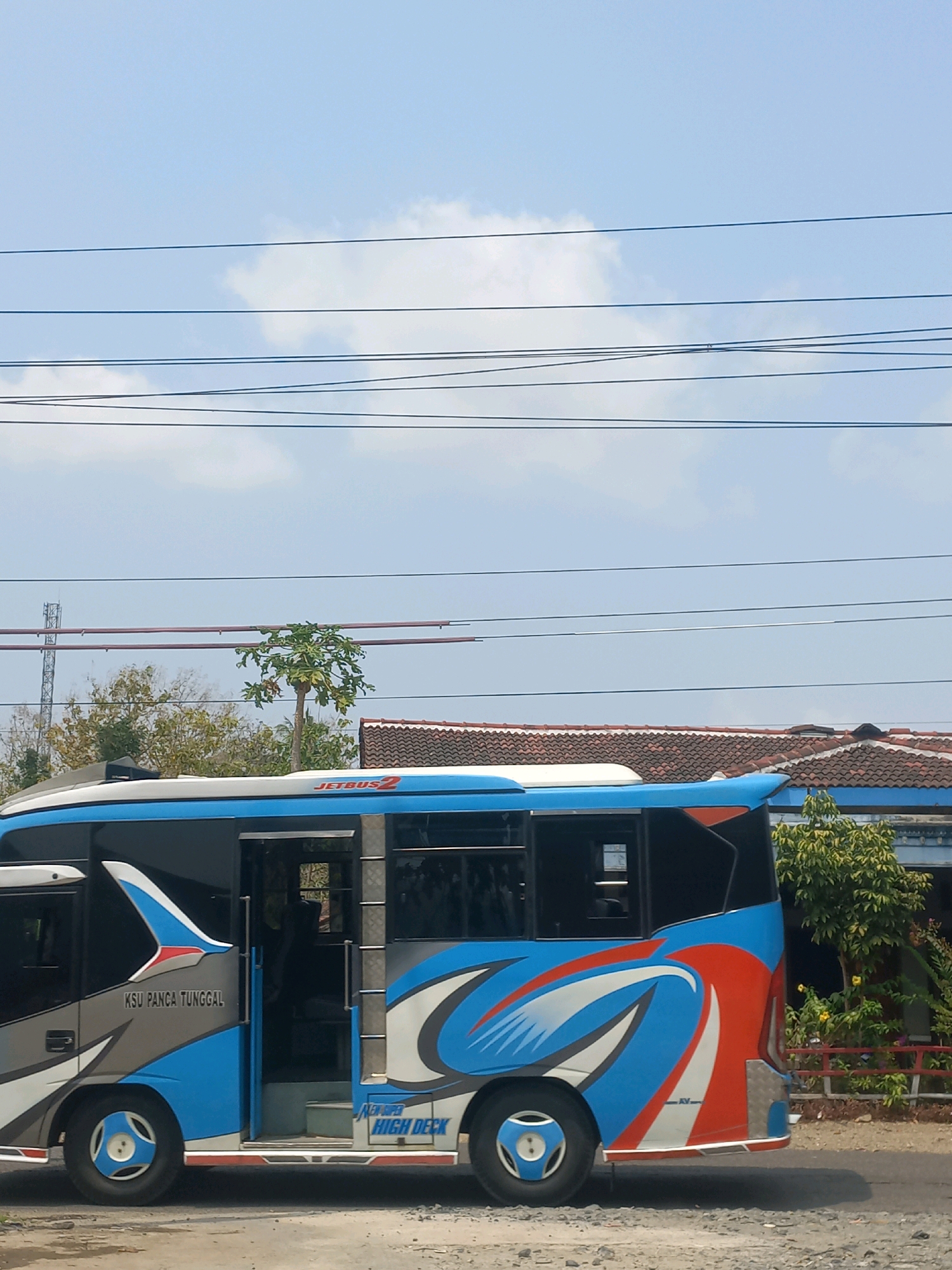 A side view of a blue and white bus is captured outdoors, likely in the city of Baturetno, Indonesia, under a bright sky with scattered clouds. The bus, identified by the text "JETBUS2" and "KSU PANCA TUNGGAL," features prominent blue, white, and red graphics. The passenger door is open, revealing a glimpse of the interior. The bus is parked on a gravel surface, with a road visible in the foreground. In the background, a building with a tiled roof and lush green trees are present. Overhead, a network of power lines crisscrosses the sky.