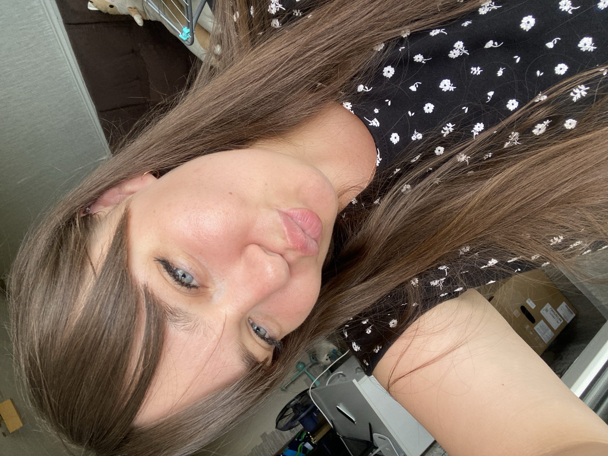 A young woman with long, brown hair and blue eyes is taking a selfie. She is wearing a black dress with a white floral pattern. Her lips are pursed, and she is looking at the camera. In the background, there is a brown couch and a stuffed animal on a drying rack. To the left, there is a desk with a computer monitor and a fan. The scene appears to be indoors, likely in a residential setting in Votkinsk, Russia.