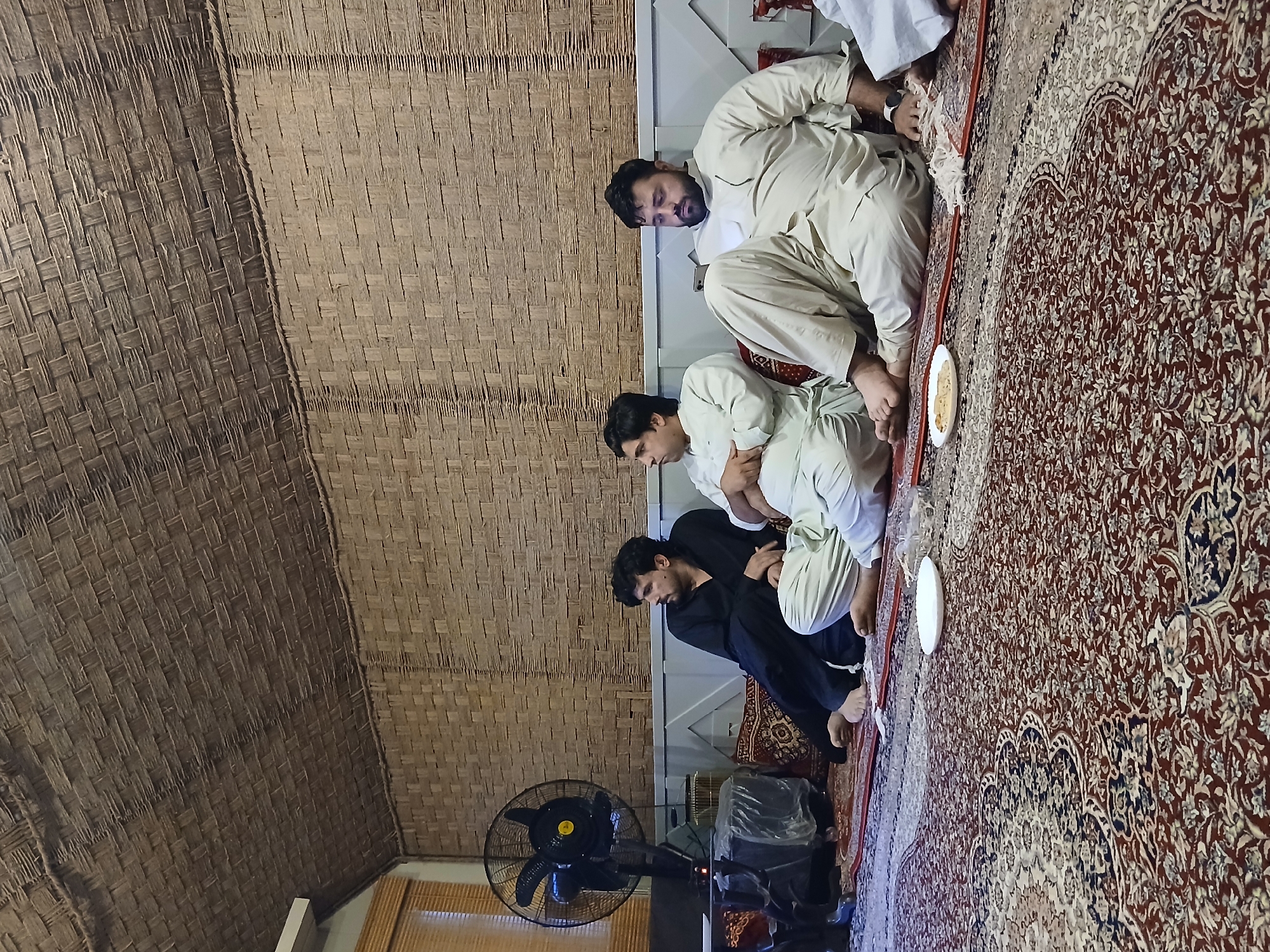 Three men are sitting on a patterned rug. One man wears a black shirt and two are wearing white shirts. They are squatting, with plates of food in front of them. A black fan stands in the corner of the room. The ceiling is woven. The location is likely in Quetta City Tehsil, Pakistan.