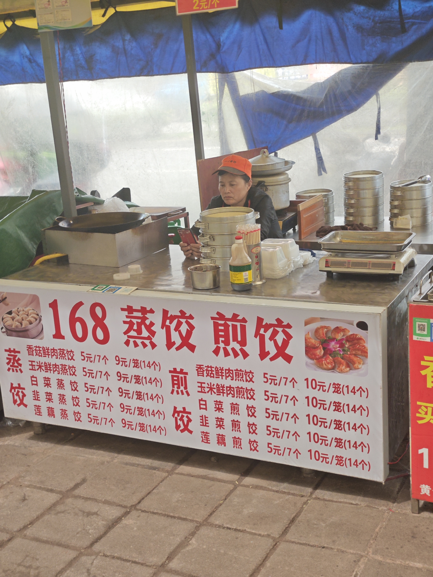 The image shows a food stall in Shapingba District, China, selling steamed and fried dumplings. A woman wearing an orange hat is standing behind the counter, looking at her phone. The sign on the counter displays the menu and prices for various types of dumplings, including mushroom and pork, corn and pork, cabbage, chives, and lotus root. The dumplings are priced at 5 yuan for 7 pieces or 9/10 yuan for 14 pieces, depending on whether they are steamed or fried. There are several steamers stacked behind the woman and a scale to her right. A green QR code is visible on the right side of the image, and the ground is paved with red bricks.