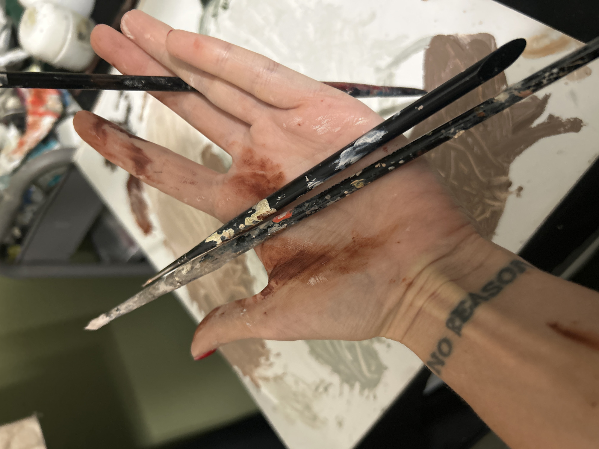 A photograph depicts a person's hand holding several paintbrushes over a surface covered in paint. The hand is also stained with paint. A tattoo reading "NO REASON" is visible on the person's wrist. The location is Cape Coral, United States of America.