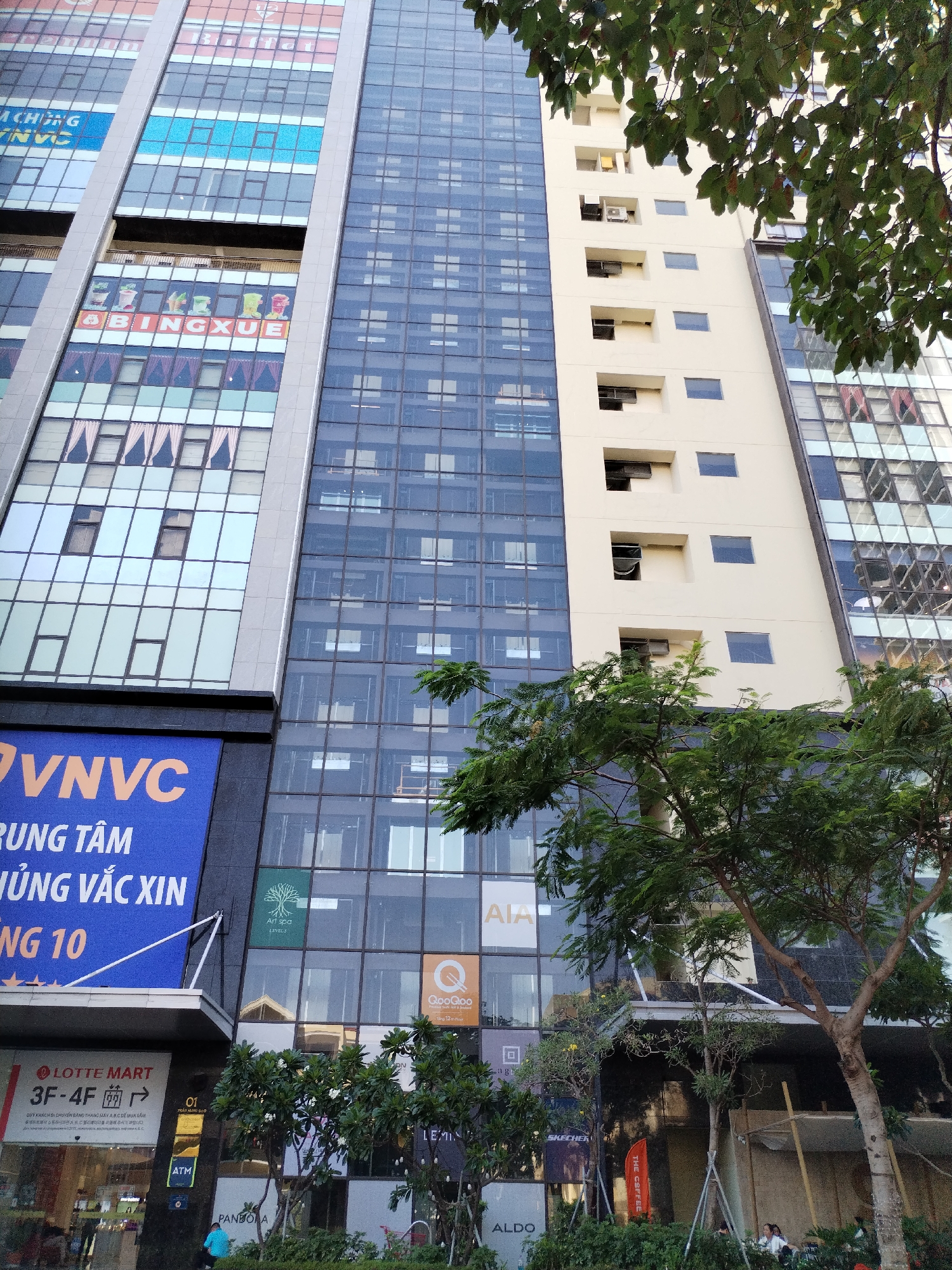 The image captures a street-level view of a mixed-use high-rise building in Nha Trang Ward, Vietnam. The building is composed of several sections with varied facades: glass curtain walls, a section with recessed windows, and commercial signage. The signage includes brands like LOTTE Mart, Art Spa, AIA, Goo Goo, Pandora, Skechers, and Aldo. A large blue sign with orange lettering reading "VNVC" and additional text in Vietnamese is prominent. Lush greenery, including trees and shrubs, partially obscures the base of the building, suggesting landscaping along the street. People are seated at tables under makeshift structures in the lower right-hand portion of the image. Overall, the scene depicts a commercial area with modern architecture and dense vegetation.