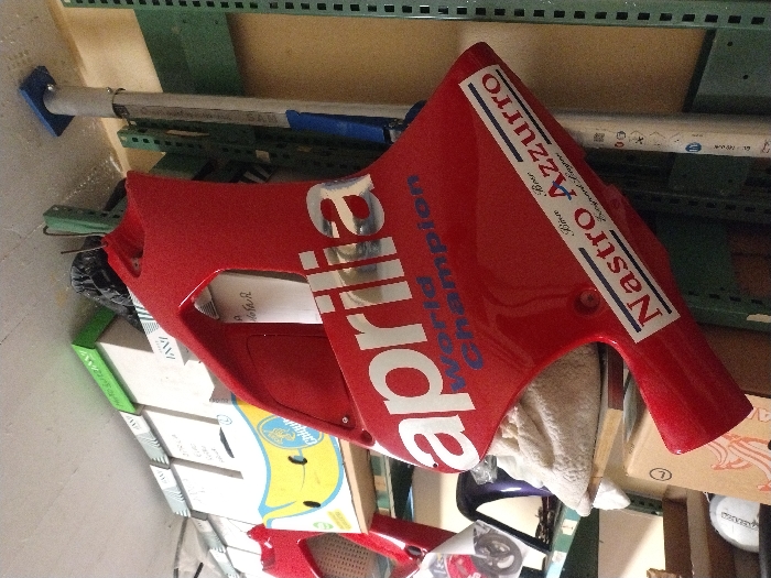 The image displays a storage area within a building in Fentange, Luxembourg. The central focus is a glossy red motorcycle fairing, likely a side panel, angled towards the viewer. It features prominent white "aprilia" lettering, "World Champion" in blue text below, and a rectangular sticker displaying "Nastro Azzurro" in bold text, with "Birra Beer" written above it.
In the background to the left, multiple cardboard boxes are stacked on shelves; one box is branded "Chiquita". Other red motorcycle components are partially visible. A light-colored, crumpled material rests behind the fairing. Further down, a white spherical object with an indistinct dark logo is partially visible.
To the right, light green metal shelving units are visible. A silver-colored adjustable support pole with a blue top, marked "SAM", extends vertically from the floor to the ceiling, adjacent to the shelving. The wall is light beige, and the ceiling is white.
No individuals are present, and no actions or interactions are depicted.