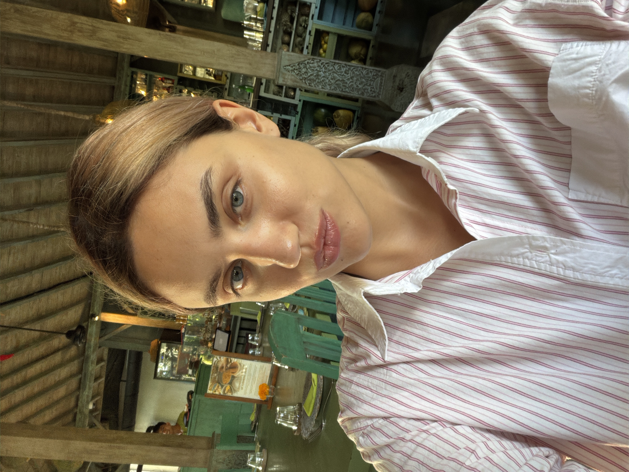 A young woman with blonde hair pulled back and light blue eyes takes a selfie at a table in what appears to be an open-air restaurant or cafe. She is wearing a pink and white striped button-down shirt. The background shows a wooden ceiling with exposed beams, a green table set for dining with glasses and placemats, and shelves filled with bottles and decorative items. A man in a green shirt is visible in the far background, working behind a counter. The lighting suggests it is daytime. The ambiance suggests a casual, tropical setting, possibly in a location like Ubud, Indonesia, known for its restaurants and cultural spaces with similar architectural styles and decor.