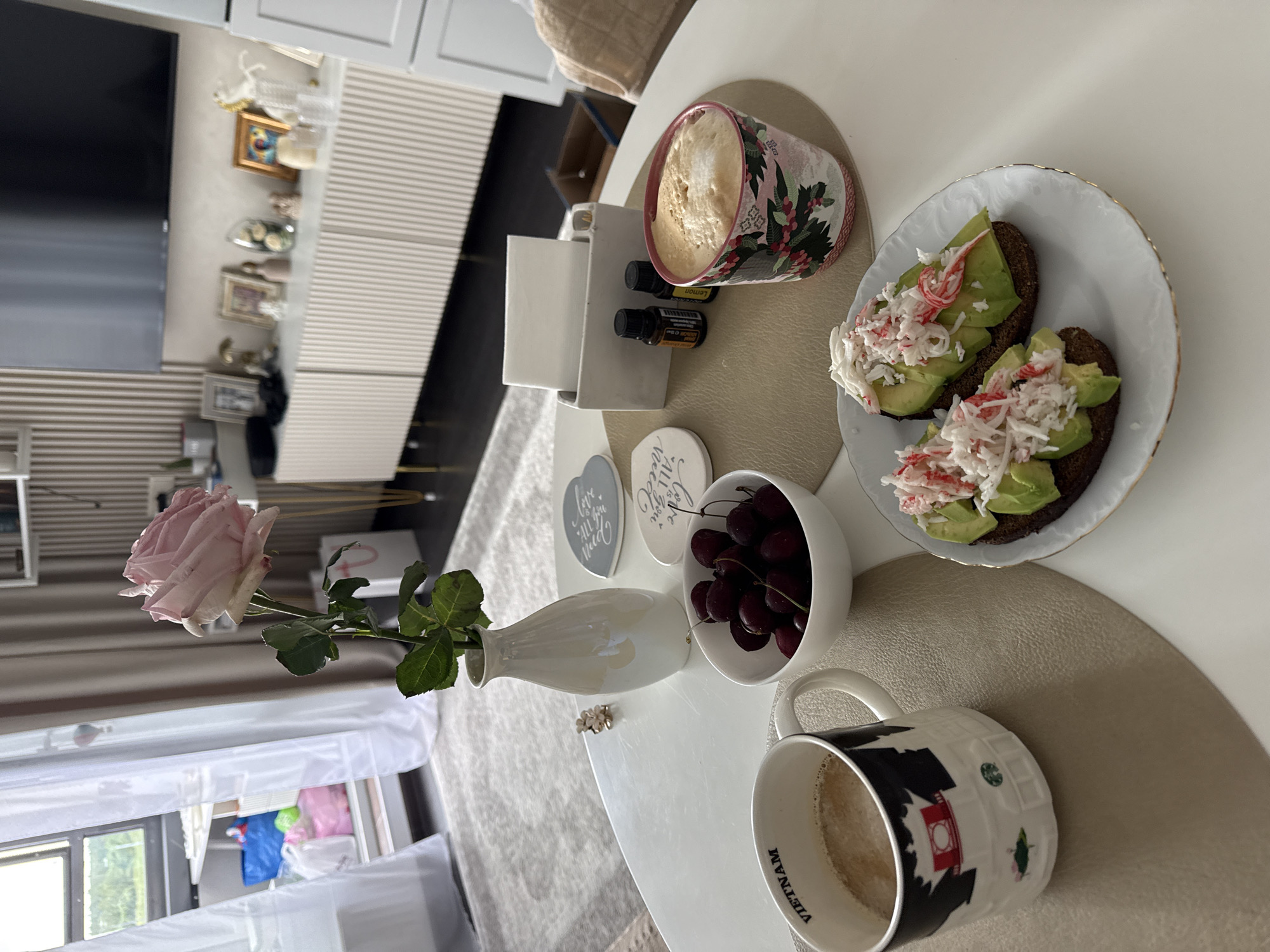 A bright, overhead shot captures a breakfast spread on a white oval table in a modern living room. In the foreground, two slices of dark toast topped with avocado and shredded crab meat sit on a white plate with a gold rim. To the left, a mug with a black and white design, reading "VIETNAM," contains coffee. Next to it, a white bowl overflows with dark cherries. A single, pale pink rose in a white vase stands in the center of the table. To the right, a ceramic mug adorned with a floral pattern holds a frothy coffee. A small white napkin holder with two small essential oil bottles sits behind it. In the background, a light-colored sectional sofa is visible, along with a media console displaying various decorative items and a television. A large window with sheer white curtains reveals a glimpse of an outdoor scene.