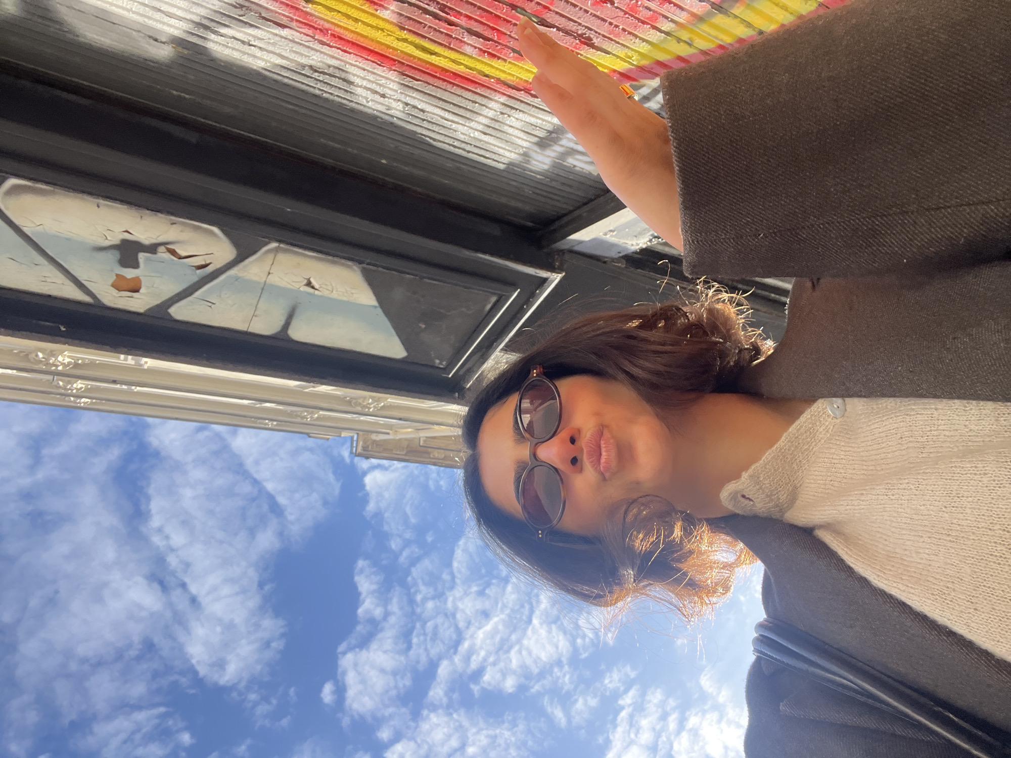 The provided image is a selfie, rotated 90 degrees counter-clockwise, capturing a young woman outdoors under a bright sky and building structure.
The woman is positioned in the lower portion of the frame (when viewed correctly oriented), wearing sunglasses and playfully pouting her lips for the camera. She has dark, wavy hair and is dressed in a light-colored (beige or off-white) knitted top beneath a dark brown or grey coat. A ring is visible on her finger, and a black bag strap rests on her shoulder.
Above and behind her, a clear blue sky with scattered white clouds is visible, indicating a bright, sunny day, likely midday or early afternoon. To the left side of the frame, ornate white classical architecture of a building can be seen, consistent with the context of Paris, France.
Directly above the woman, the underside of a building element, possibly an awning or balcony, is prominent. This structure features dark-framed sections with severely peeling, distressed paint that shows light blue and brownish-orange tones. Further along this structure, there are horizontal ribbed surfaces, some brightly painted with distinct red and yellow stripes.
The scene conveys a casual and cheerful mood. No visible text is discernible in the image.