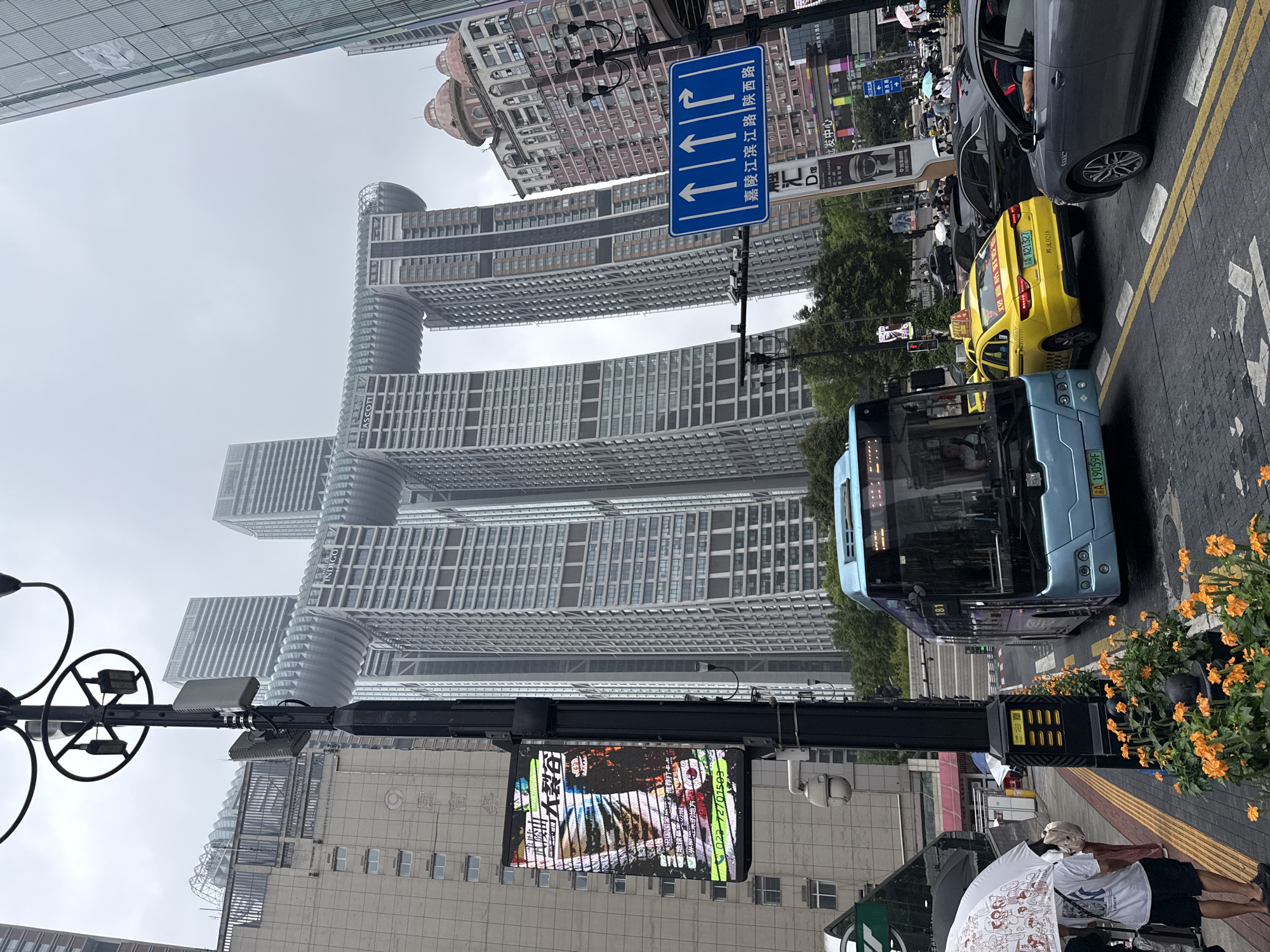 In Yuzhong District, China, on an overcast day, a busy street scene unfolds. The focal point is the distinctive Raffles City Chongqing complex, a collection of interconnected skyscrapers that form a horizontal element resembling a ship or a dragon. In the foreground, a light blue public bus with the number "181" displayed is stopped at a traffic light. Beside it, a yellow taxi bearing the license plate "AA A21327" is also waiting. Several other vehicles, including a dark grey car, are visible in the traffic.
On the sidewalk to the left, a person holding a white umbrella with a cartoon print stands. A lamppost with antennas and what appears to be a surveillance camera is prominent in the mid-ground. A digital advertisement billboard is mounted on the lamppost, displaying colorful graphics and text. Orange flowers are planted in raised beds along the sidewalk. The environment is characterized by modern, tall buildings, suggesting an urban setting. A blue traffic sign with white arrows and Chinese text indicates lane directions. The overall atmosphere suggests a typical day in a bustling Chinese metropolis.