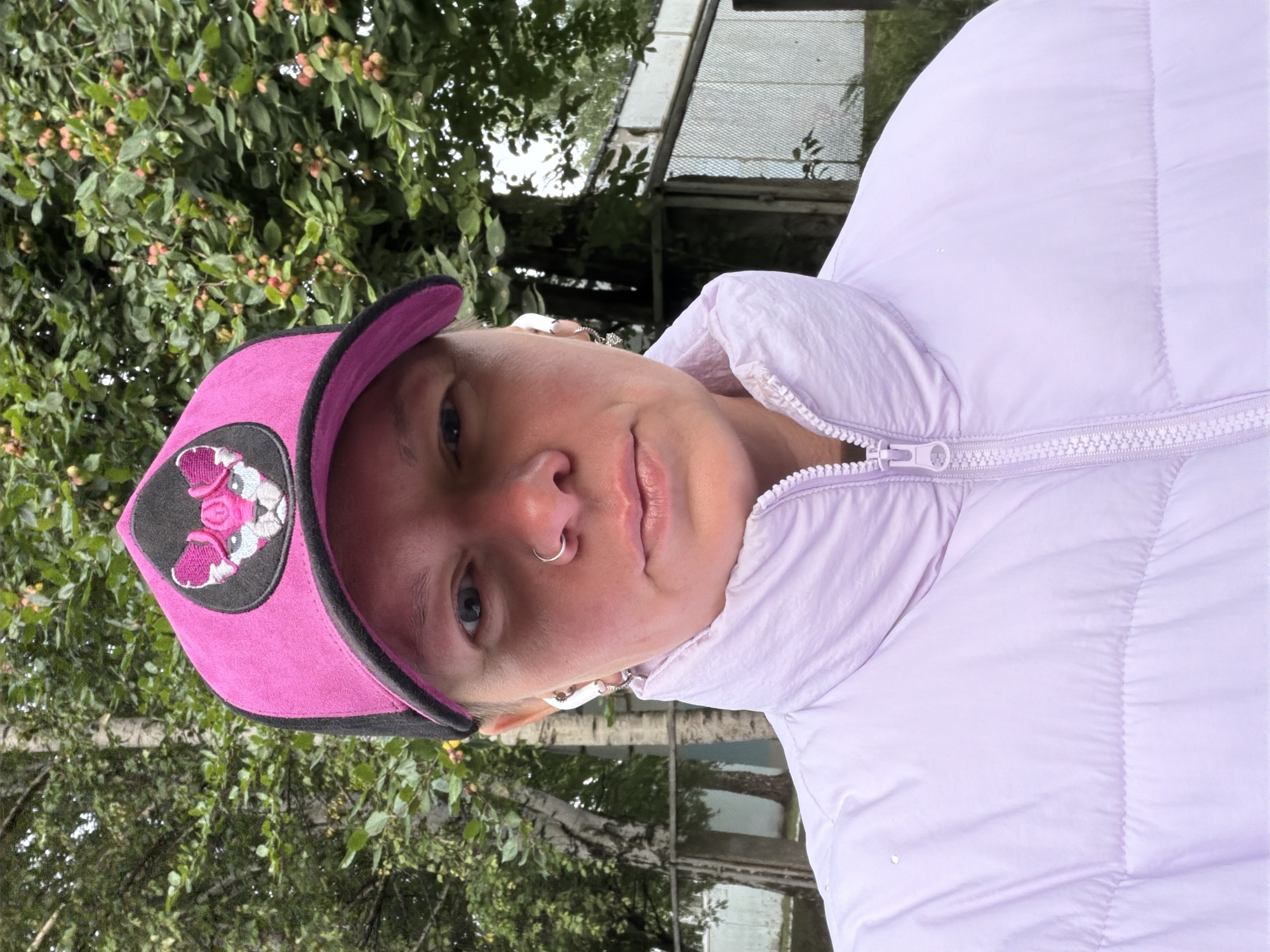 A person wearing a pink baseball cap with a black brim and a patch depicting a pink animal's face, and a light purple puffer jacket, is shown in a close-up selfie outdoors. The person has a nose ring and is wearing white wireless earbuds. The background consists of green trees with small red fruits, a birch tree, and a chain-link fence. The image appears to be taken in Chaikovsky, Russia, judging by the presence of birch trees and a rustic, possibly dacha-like environment. The lighting suggests an overcast day.