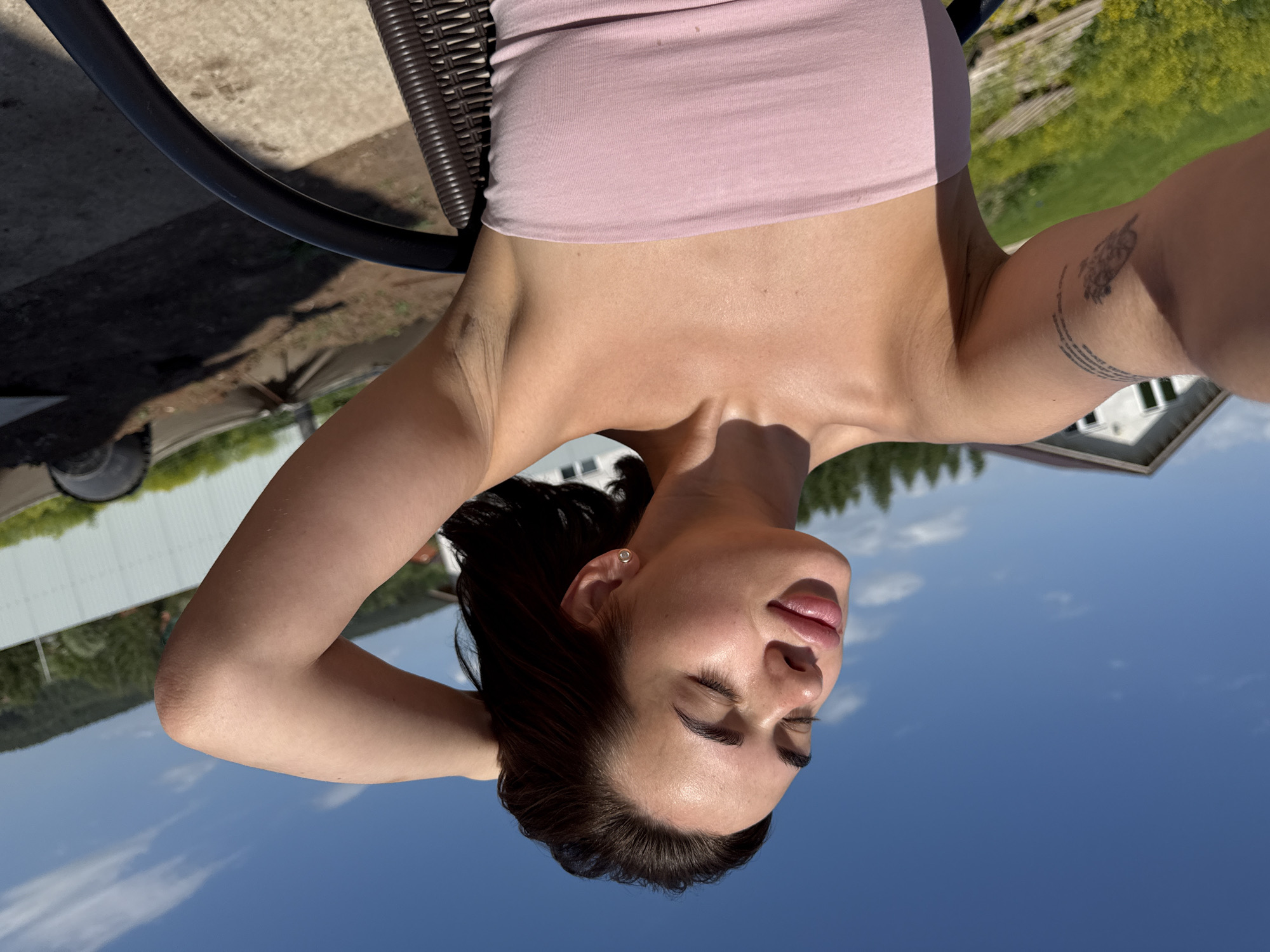 A woman is lying on a woven chair outdoors on a sunny day with a blue sky visible above. She is wearing a pink strapless top, and her dark hair is pulled back. Her eyes are closed, and she appears to be relaxing. A tattoo is visible on her upper arm. The background includes a building and green foliage. The location is likely in a sunny outdoor area, possibly in Лобаново, Russia.