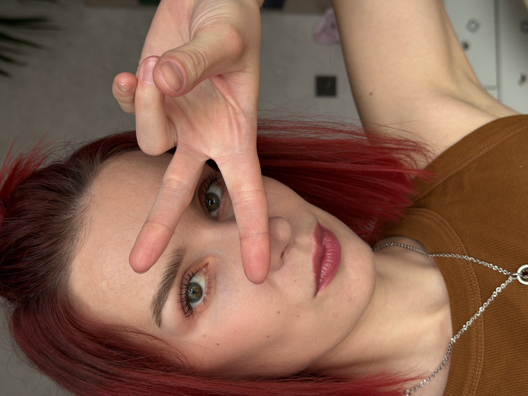 A close-up, slightly overhead portrait depicts a young woman with vibrant red hair, fair skin, and striking green eyes, looking directly into the camera. Her head is tilted, giving the image a dynamic, rotated perspective. She makes a "peace" sign (V-sign) with her right hand, with her index and middle fingers partially obscuring her nose and forehead. Her eyelashes are enhanced with mascara, and her lips are painted with a pink lipstick. She wears a brown ribbed top and a simple silver chain necklace. Her expression is neutral but direct. The background is softly blurred, suggesting an indoor setting, possibly a room with a light-colored surface to the right and faint plant-like shapes to the left. The image is rich in detail, highlighting her facial features, hair color, and hand gesture. There is no visible text in the image. The location context is Chaikovsky, Russia.
