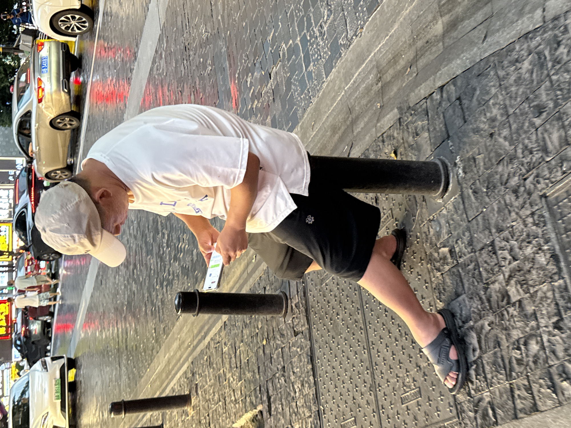 A man wearing a white t-shirt, black shorts, and a light-colored baseball cap sits on a black bollard on a wet, paved city street in Yuzhong District, China. He is holding and looking at a smartphone. In the background, several cars are visible on the street, including a white sedan on the left, a silver sedan in the middle, and a white car on the right. The street appears to be illuminated by streetlights and reflections from the wet surface.
