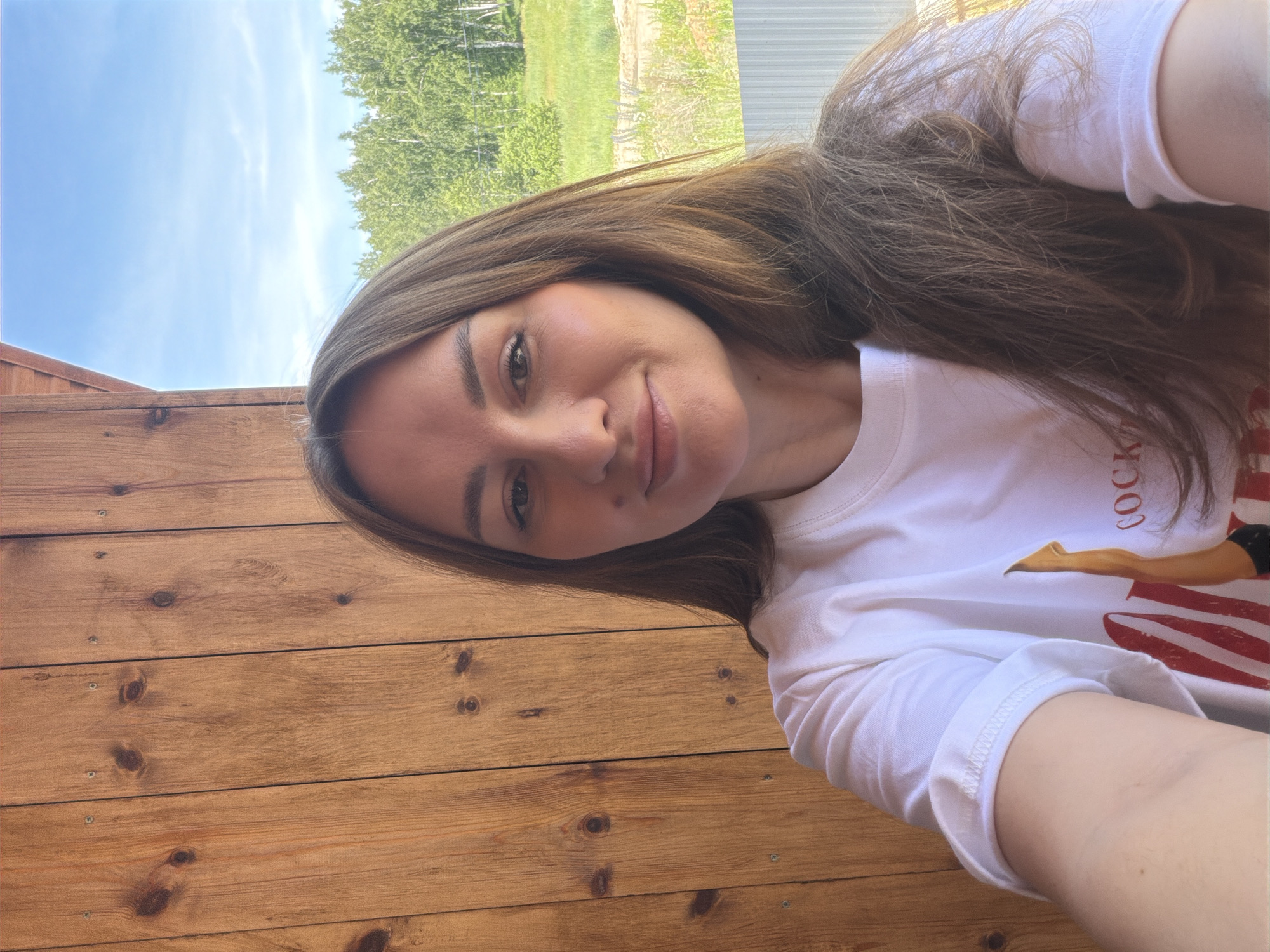 The image shows a selfie of a woman with long brown hair, wearing a white t-shirt with a graphic print. She is standing in front of a wooden wall. In the background, there is a green landscape with trees under a bright blue sky. The location is identified as Malinovka, Russia.