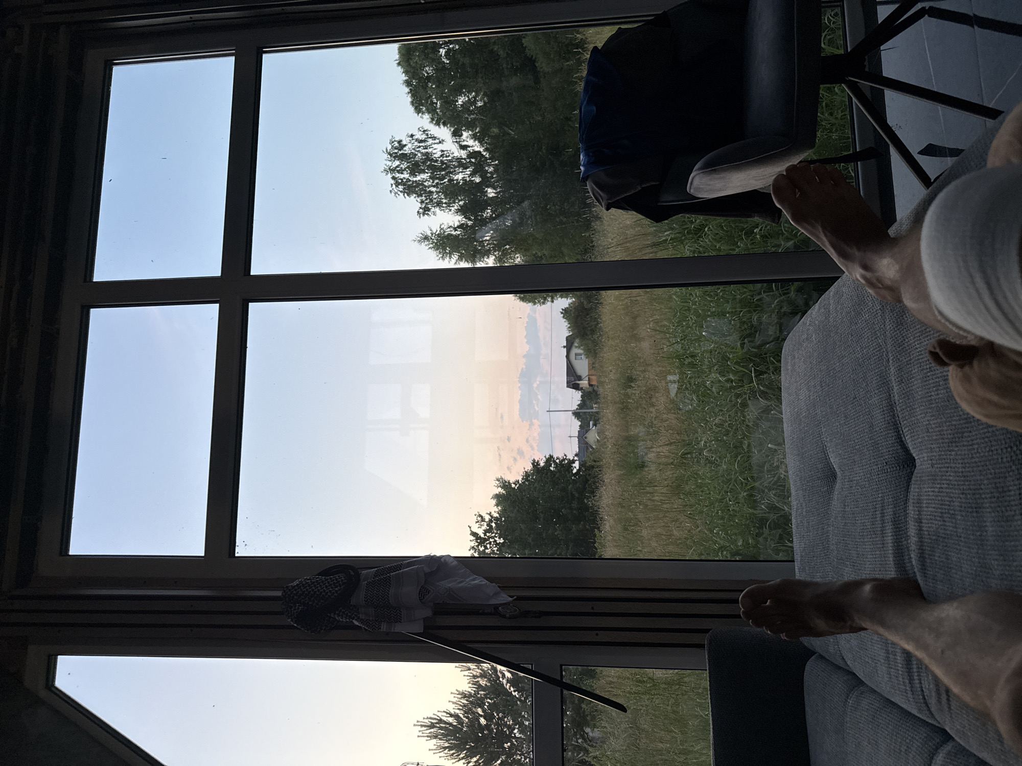This image depicts a person's legs resting on a sofa, looking out of a large window. The window frames a rural landscape with tall grass, trees, and houses in the distance, suggesting the location is the city of Vol'noe, Russia. The sky is a pale blue with hints of pink and orange, indicating either sunrise or sunset. A chair with clothing draped over it is visible to the right of the frame. A patterned cloth and a belt hang from the window frame on the left. The overall scene conveys a sense of relaxation and domesticity.