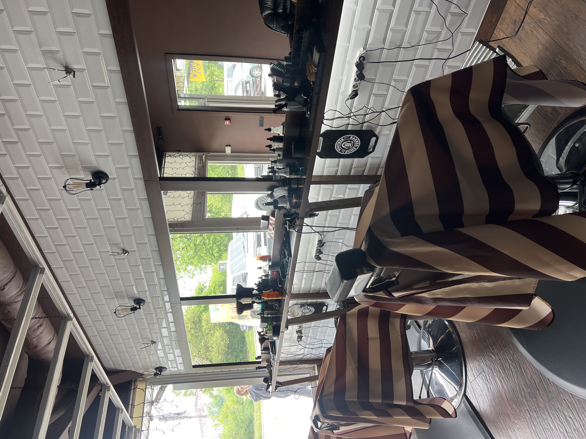 The image depicts the interior of a barbershop. Two barber chairs are draped with striped barber capes. The background includes barber stations positioned along a large window, featuring various barber supplies and tools. Outside the window is a view of what appears to be a street with trees and vehicles. The location is Ternopil, Ukraine, based on the Cyrillic lettering visible on some of the product labels. The scene is well-lit and appears to be a working barbershop with at least one customer getting a haircut.