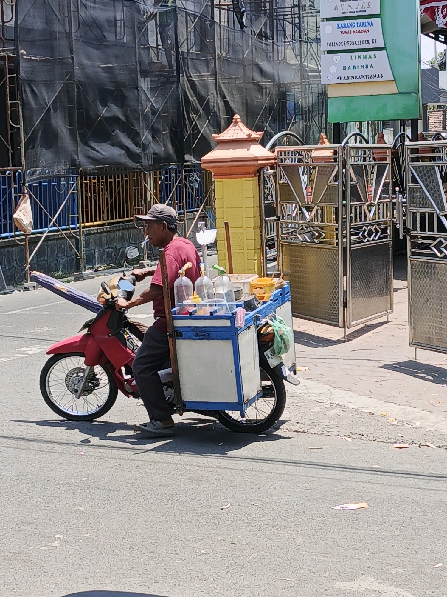 A man is riding a red motorcycle with a cart attached to the back, selling drinks. The cart contains multiple bottles of liquid and some bowls of food. The man is wearing a red shirt, dark pants, and a baseball cap, and is smoking a cigarette. He is in a street in Ketegan, Indonesia, with buildings and scaffolding in the background. The street is paved and there is a sidewalk next to it. The day is sunny.