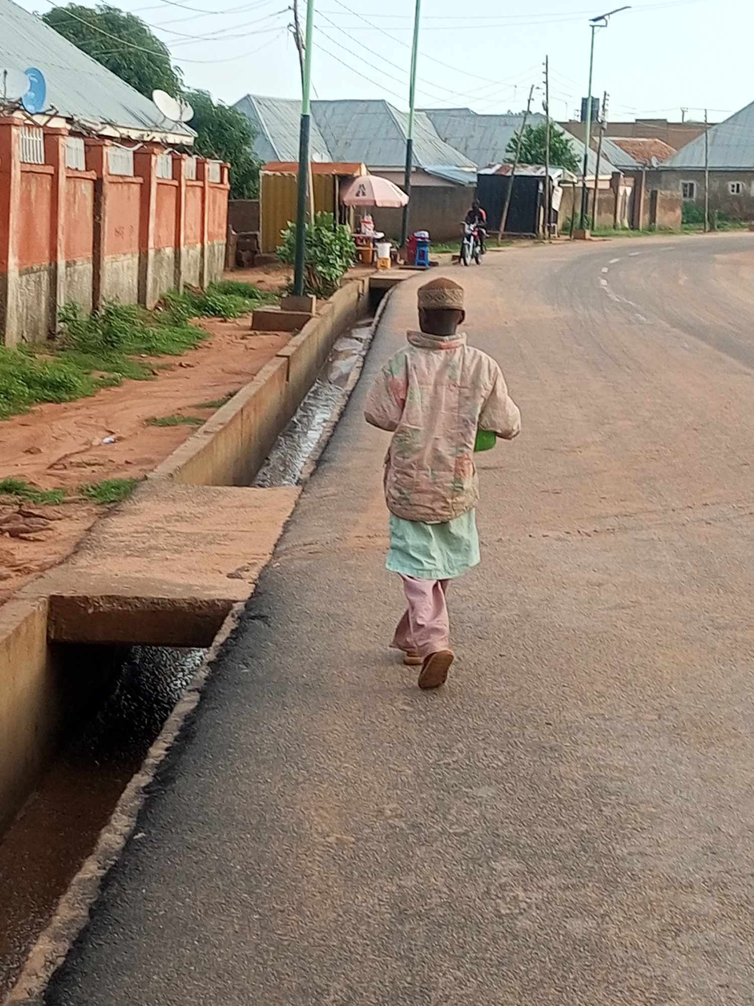 A single person walks away from the camera on a paved road in Tempure, Nigeria. The individual is wearing a light-colored, patterned jacket over a light green skirt and pink trousers. They are also wearing a patterned cap and light brown shoes, and carrying a green object in their right hand. To the left of the road is a concrete drainage ditch with water flowing through it. Beyond the ditch, there is a red and gray wall, grass, and a building with a corrugated metal roof. To the right of the road, there are more buildings with corrugated metal roofs and a person on a motorcycle in the distance. The sky is clear and bright.