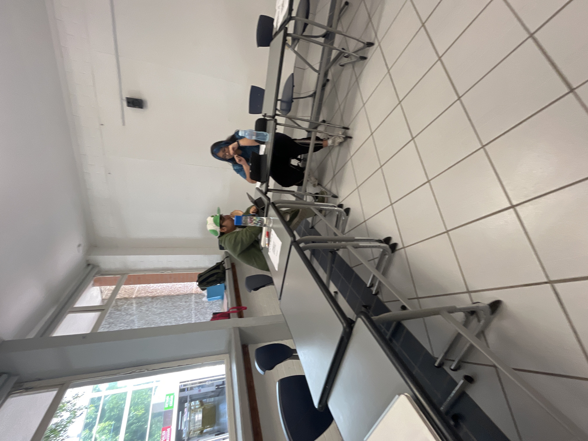 The image shows the interior of a room, possibly a classroom or study area, in what is suggested to be Puebla, Mexico. The room features rows of tables and chairs aligned in a parallel arrangement. Two people are visible at one of the tables. The first person, closer to the camera, appears to be male wearing a green shirt, and is sitting at the table with items on top. The second person is a female sitting at the table and drinking water from a bottle. Windows can be seen in the background of the room showing trees and a sky outside.