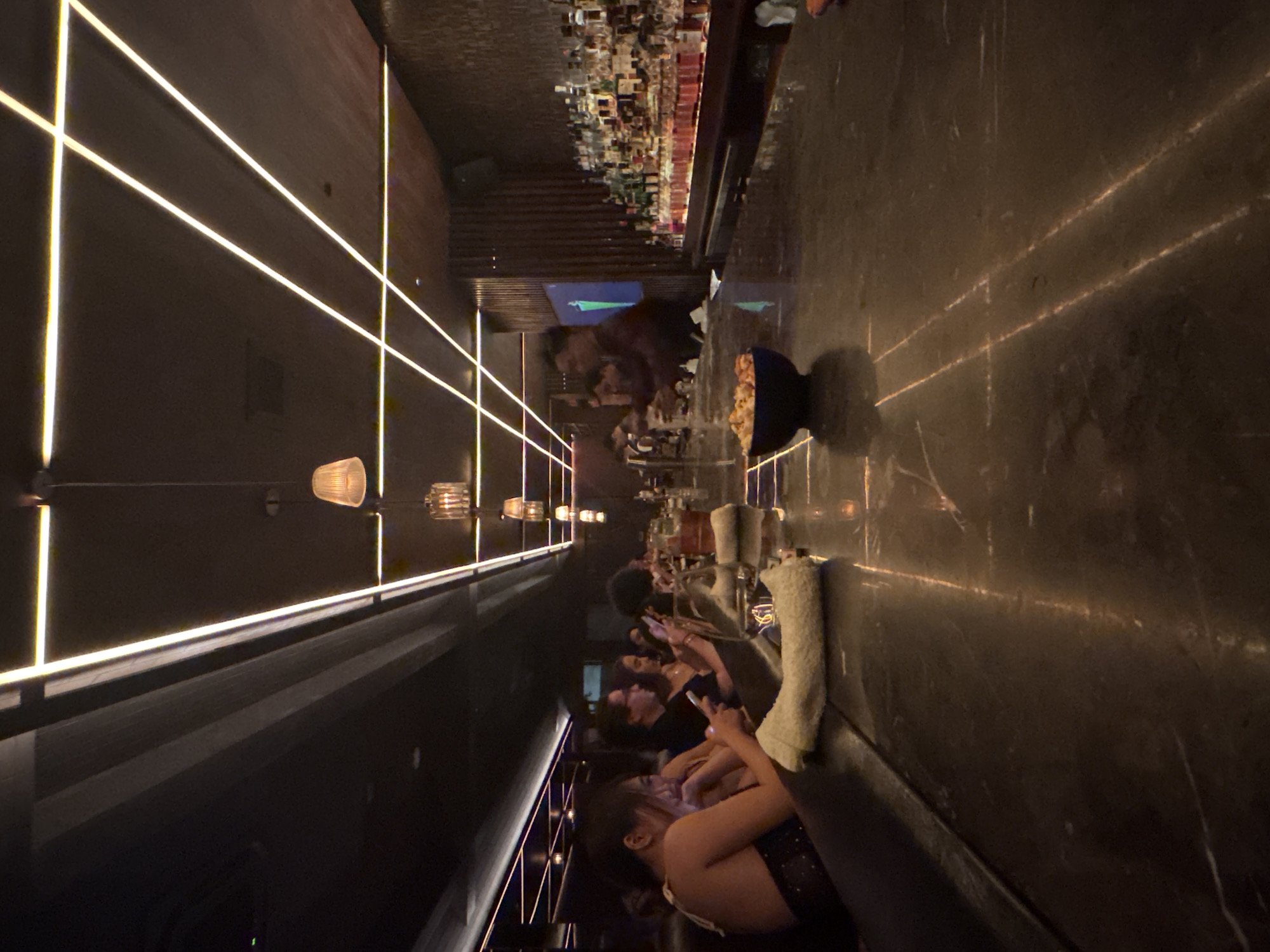 The image depicts a bar in North Kuta, Indonesia. A line of people, seemingly customers, are seated at the bar. Most are looking at their phones. The bar itself is long, with a wide countertop. A shelf of bottles runs along the back wall of the bar. There is decorative lighting on the wall behind the bar, consisting of parallel lines of light. Overhead are various hanging light fixtures. A bowl of popcorn is visible on the bar.
