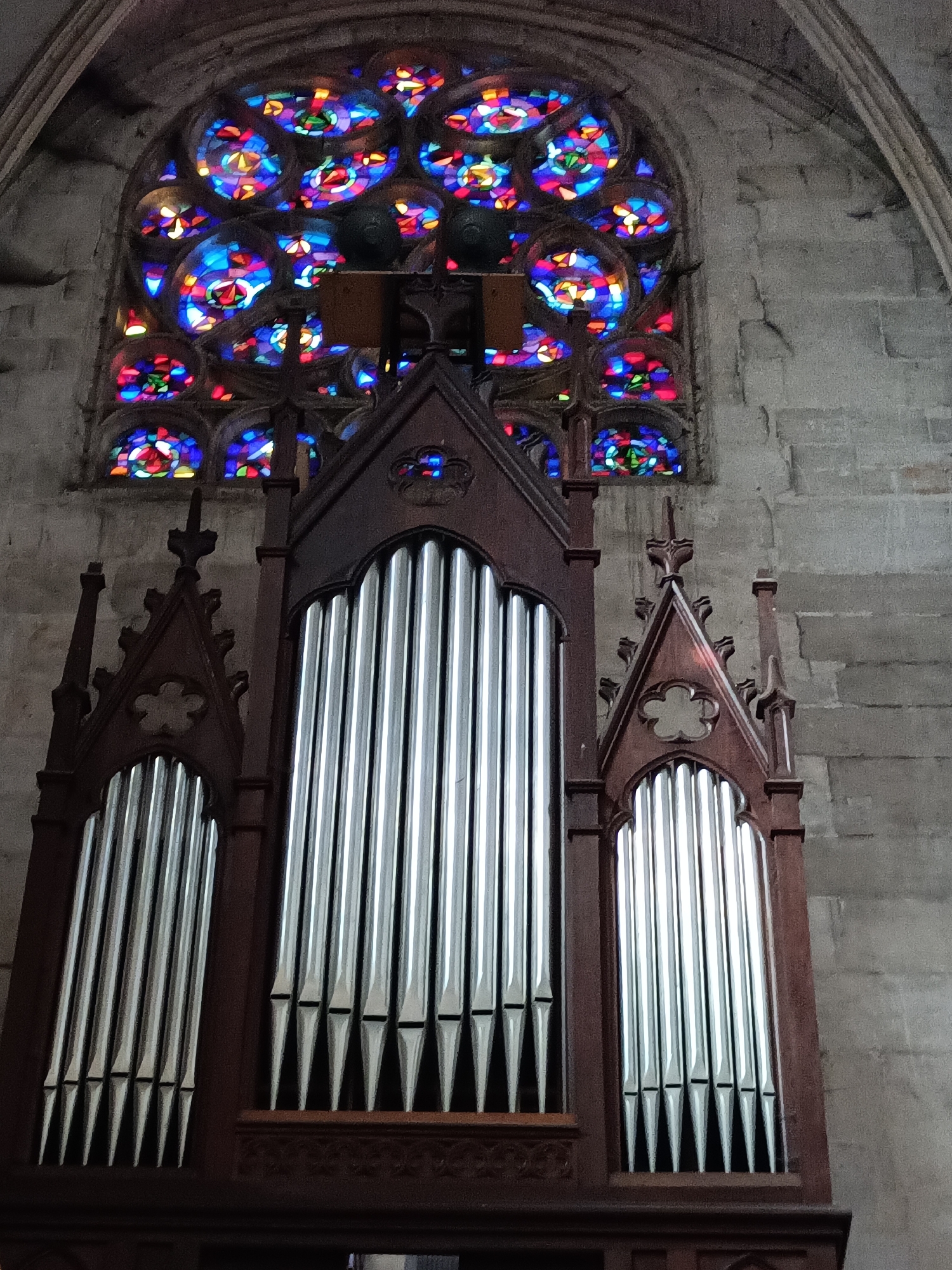The image shows a pipe organ positioned in front of a large, circular stained-glass window in a stone building. The organ has a dark wooden casing and numerous silver-toned pipes of varying lengths. The stained-glass window above is highly ornate, featuring intricate geometric patterns and a vibrant array of colors, including blues, reds, purples, and yellows, illuminated by light passing through it. The stone walls of the building are visible to the right and above the window, composed of large, rough-hewn blocks. The organ and the window are located within the church of Saint-Thomas-Apôtre in Le Neubourg, France.