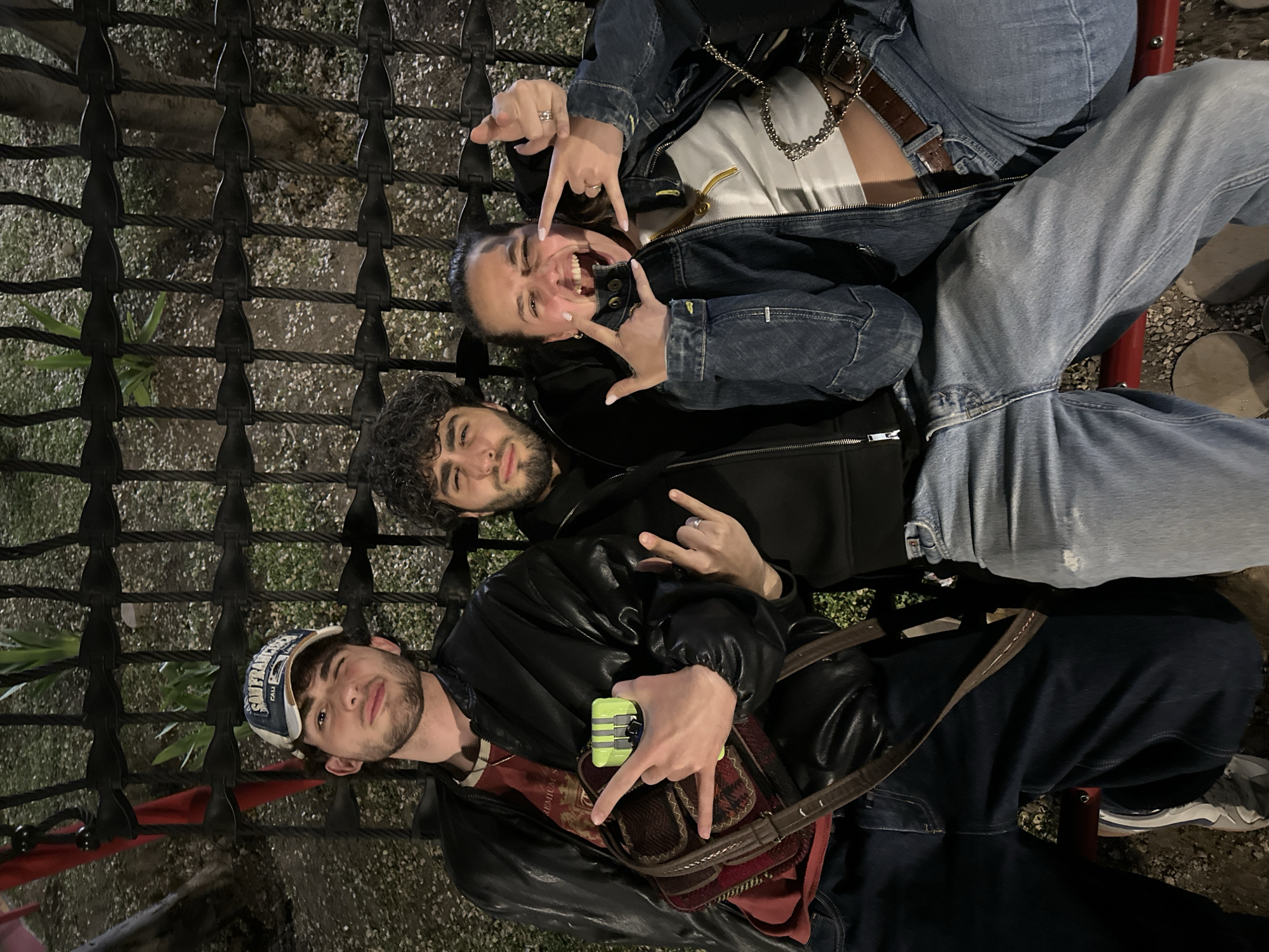 Three young adults pose on a seating structure made of black rope and metal. The woman on the top wears a jean jacket and jeans, grinning widely, and is throwing a "rock on" sign with both hands. The man in the middle has curly dark hair and a beard, wearing a black hoodie, also displaying the "rock on" sign. The man at the bottom wears a leather jacket and a baseball cap and is holding a small object, also making the "rock on" sign. They appear to be outside at night. The background reveals what looks like ground cover and possibly some greenery. This location is possibly in Yerevan, Armenia.