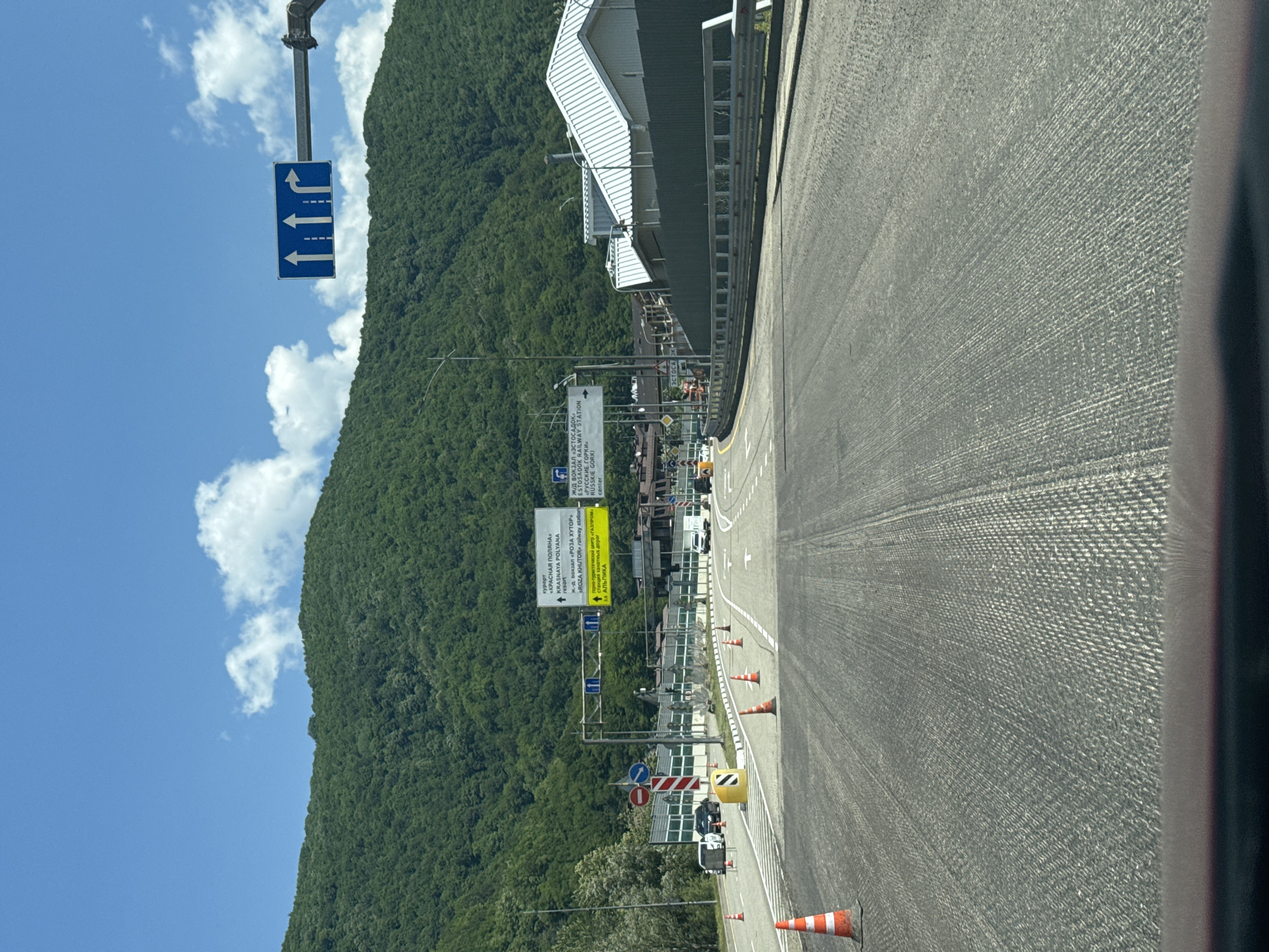 The image depicts a road leading towards a mountainous area, likely in Estosadok, Russia, given the signage. The road surface in the foreground shows recent resurfacing work with a patterned texture. Several orange traffic cones are placed along the sides of the road, indicating ongoing or recent road construction or maintenance. In the distance, there are traffic signs directing vehicles, including a blue sign with three upward arrows and a right-turn arrow, and yellow directional signs in Russian. The road curves to the right and appears to lead to a checkpoint or a populated area with buildings visible. The environment is characterized by a lush green mountain covered in trees under a clear blue sky with scattered clouds. There are no people clearly visible in the scene, and the focus is on the road infrastructure and the natural landscape.