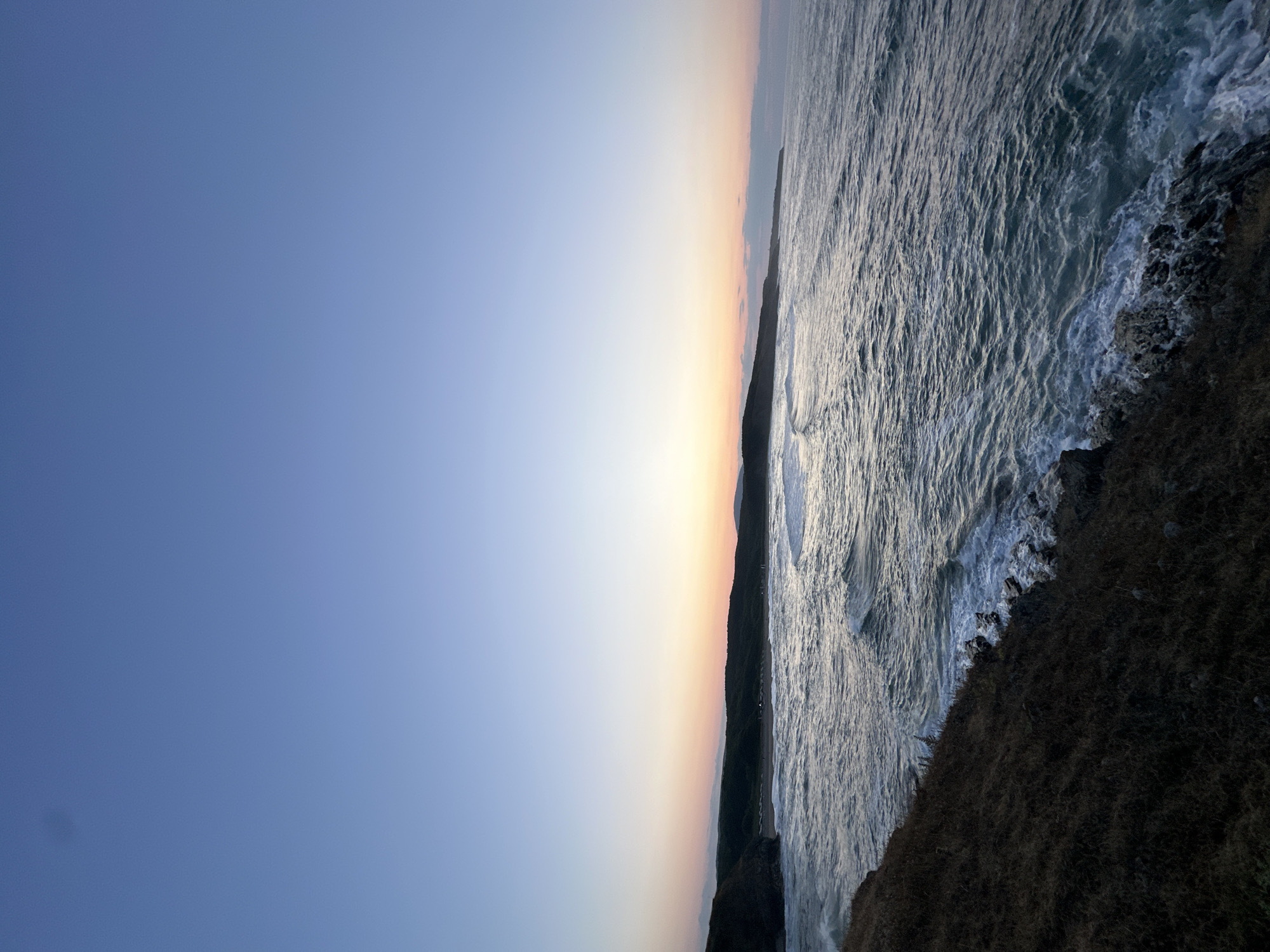 A wide-angle, low-angle shot captures the Black Sea coast at Sinemorets, Bulgaria, during twilight. The sky transitions from a deep blue overhead to a soft gradient of pink and orange near the horizon, hinting at the recent sunset. The sea is active with moderate waves, their crests illuminated by the fading light, creating a textured pattern across the water's surface. Rocky outcrops and a grassy incline form the foreground, leading the eye towards the calmer waters and the distant, rolling hills of the coastline. The beach area, visible in the mid-ground, appears to have scattered beach chairs or umbrellas. No people are visible. The overall scene conveys a sense of peaceful solitude and the raw beauty of a coastal landscape at dusk.