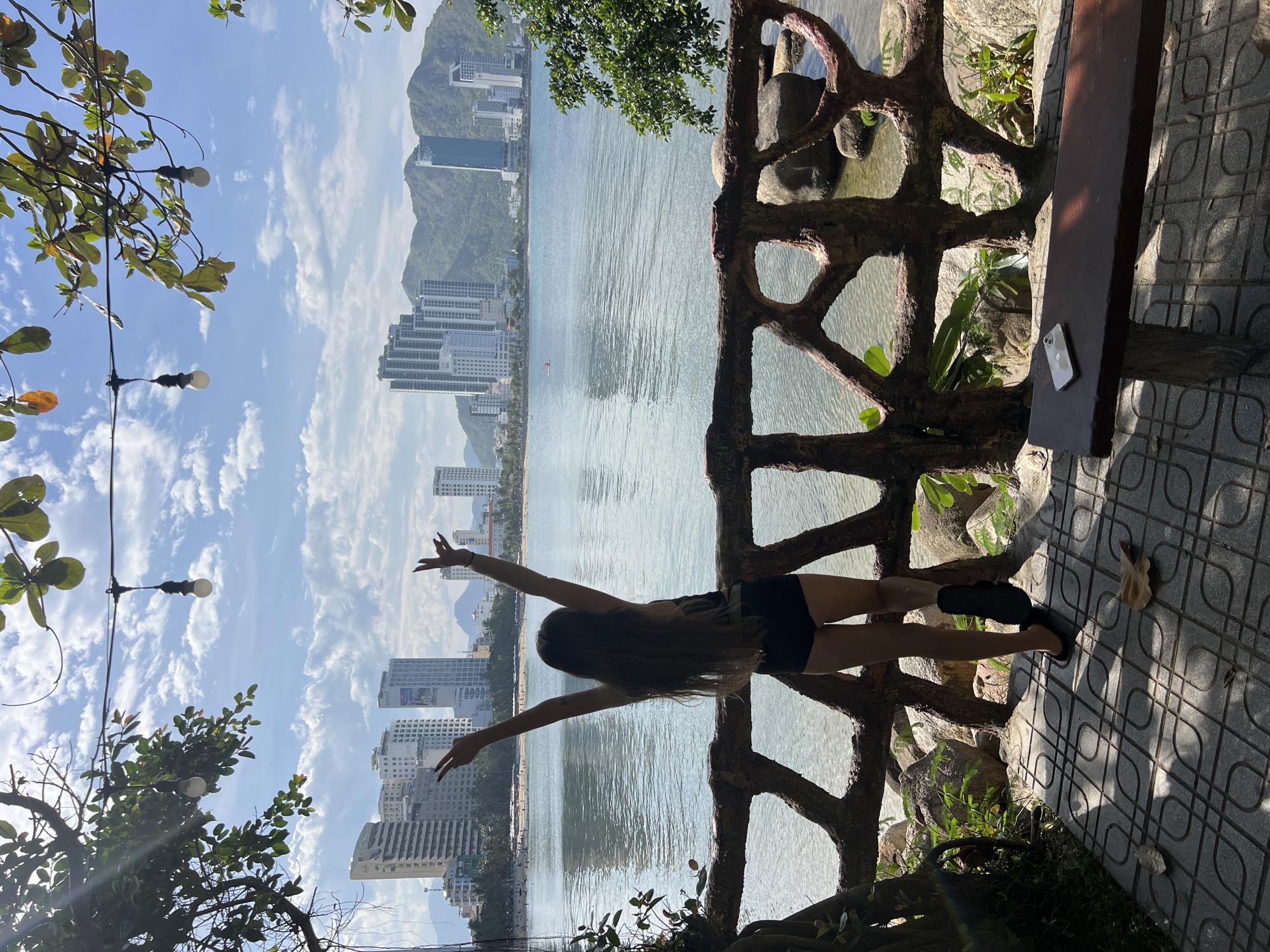 A full-shot, eye-level, outdoor, daytime photograph shows a young woman with long, dark hair wearing black shorts and a black top, standing with her back to the camera, facing a body of water. Her arms are raised in the air, with her hands in a V-shape, as if cheering or celebrating. She is standing behind a rustic, wooden railing with an organic, branch-like design. The railing encloses a small patio area paved with decorative square tiles. In the foreground, to the right of the woman, is a wooden bench with a smartphone resting on it.
Beyond the railing and the water, a city skyline is visible. Numerous high-rise buildings, predominantly white and glass, are clustered along the coastline, with a mountain range rising behind them. The water appears calm, reflecting the blue sky dotted with white clouds. Lush green foliage from trees overhangs the scene from the top and sides. The lighting suggests a bright, sunny day with some shadows cast on the patio tiles. The scene appears to be a scenic viewpoint overlooking the bay of Nha Trang, Vietnam, possibly in the Bac Nha Trang Ward area.