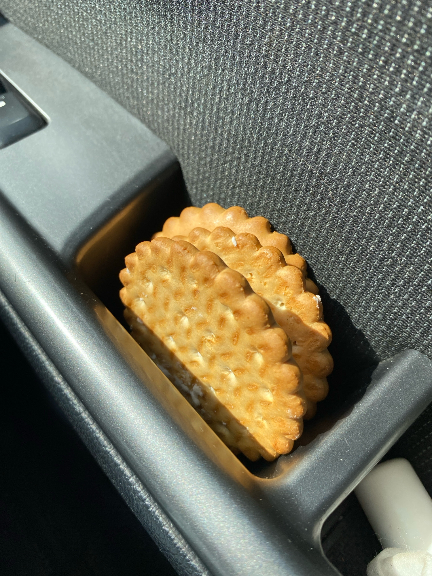 Three cream-filled crackers are stacked in a gray plastic cup holder, likely inside a vehicle. The crackers have a scalloped edge and are golden brown in color. The cup holder is positioned next to a textured gray fabric surface, possibly a car seat. Sunlight casts a shadow on the crackers from the left side of the frame. The scene is observed from a slightly elevated, close-up perspective.