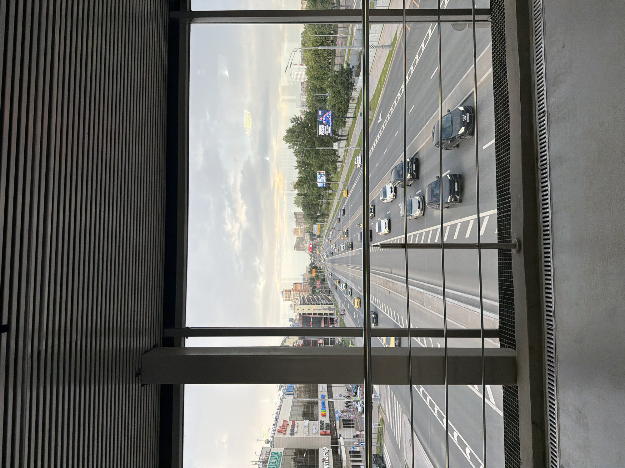 This is a medium shot, eye-level, indoor photograph taken through a large glass window. The scene depicts a multi-lane highway in an urban environment, viewed from what appears to be an elevated walkway or building. The highway is busy with traffic, including cars, taxis, and possibly buses. The cars are of various makes and models, and their headlights are on, suggesting it is dusk or early evening. The highway is lined with trees on one side, and buildings and commercial establishments are visible in the background. The sky is overcast with a gradient of light, indicating a twilight setting. In the foreground, a concrete walkway with metal grates and a metal railing are visible, suggesting a pedestrian area. The overall impression is that of a modern city with significant vehicular activity.