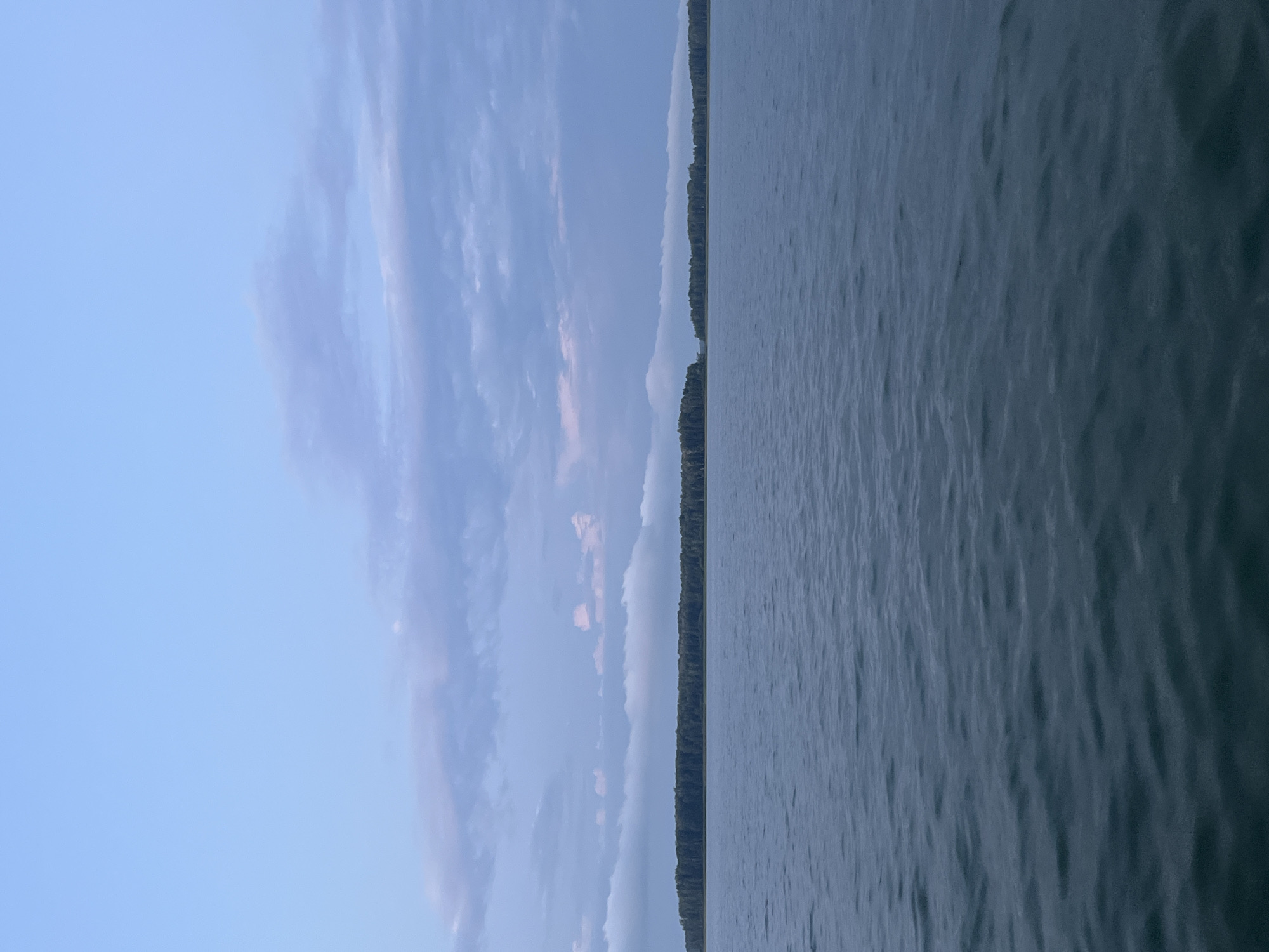 A wide body of calm, dark water stretches across the lower half of the frame, with gentle ripples disturbing its surface. In the distance, a narrow, tree-covered island forms a dark, horizontal band across the horizon. Above the island, a strip of pale white clouds is visible, paralleling the landmass. The upper half of the image is dominated by a serene sky of pale blue, transitioning to a softer, lilac hue near the horizon. Scattered, wispy clouds drift across the sky, tinged with subtle shades of pink and gray, suggesting the soft light of dawn or dusk. The scene depicts a tranquil natural landscape.